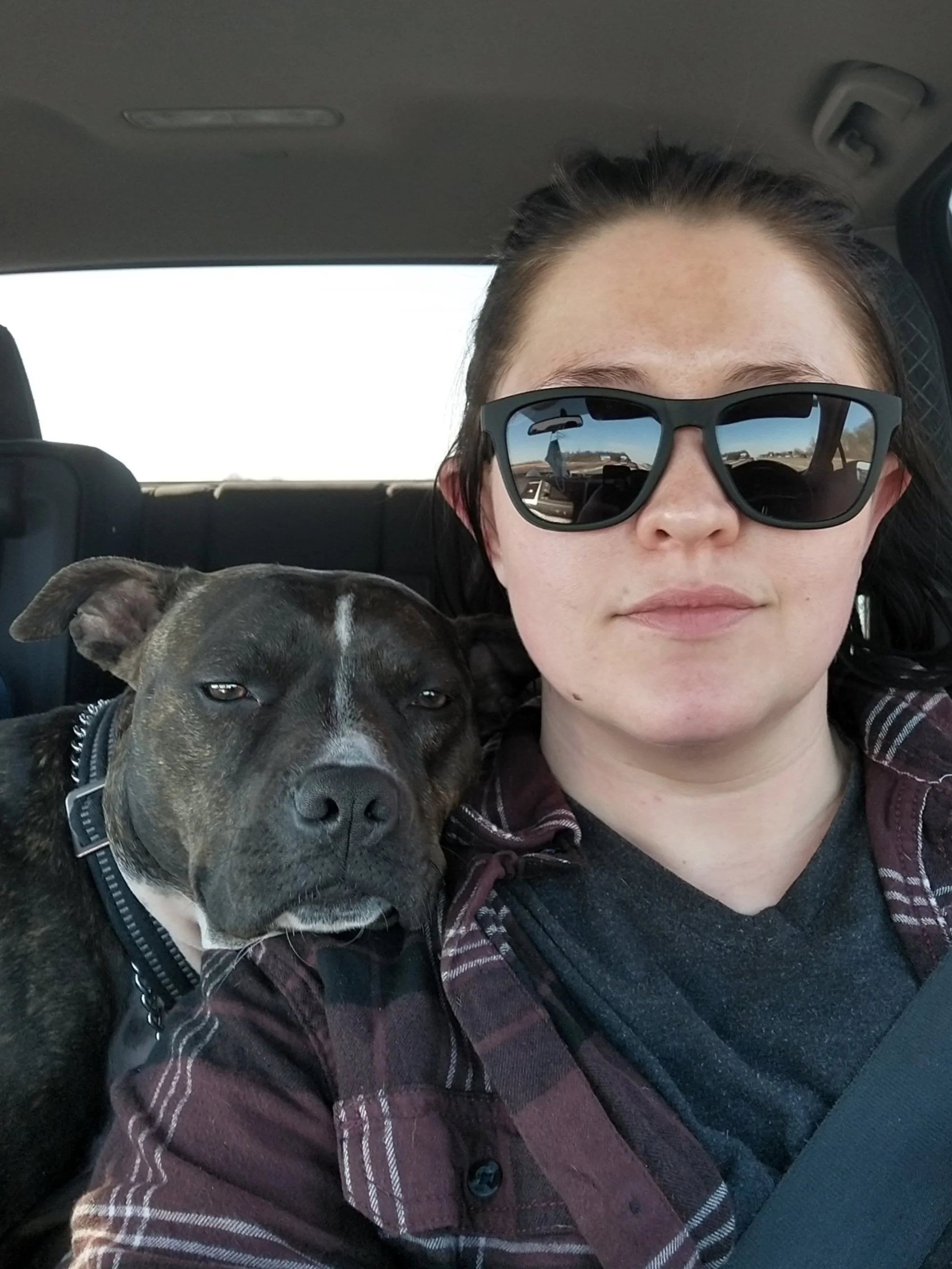 white fem person sitting in a car. they're wearing sunglasses and red flannel with their dog resting her head on their shoulder.