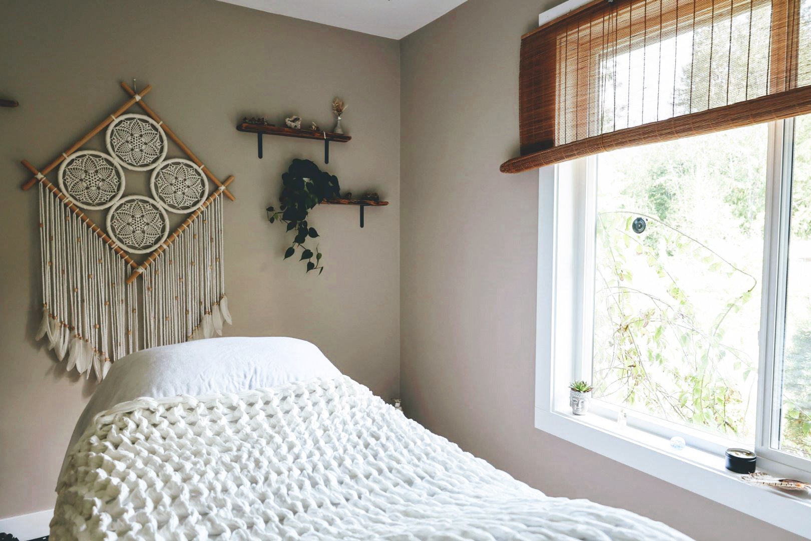 Cozy bedroom with a white knitted blanket on the bed, a macrame wall hanging with circular patterns, floating shelves with decor items, and a window with bamboo blinds.