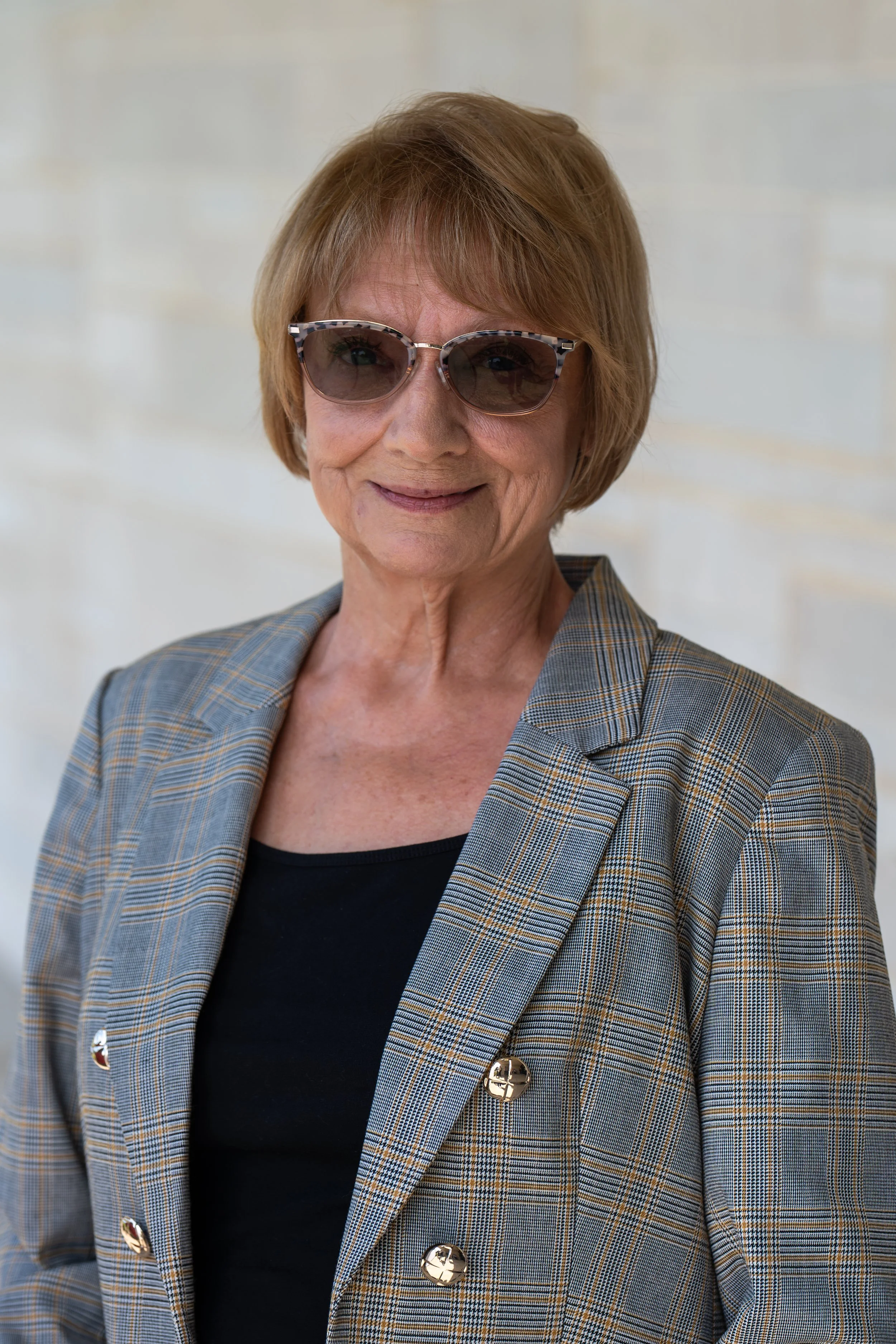 Close-up photo of an elderly woman with short red hair, wearing glasses, smiling, dressed in a white sweater, against a neutral background.