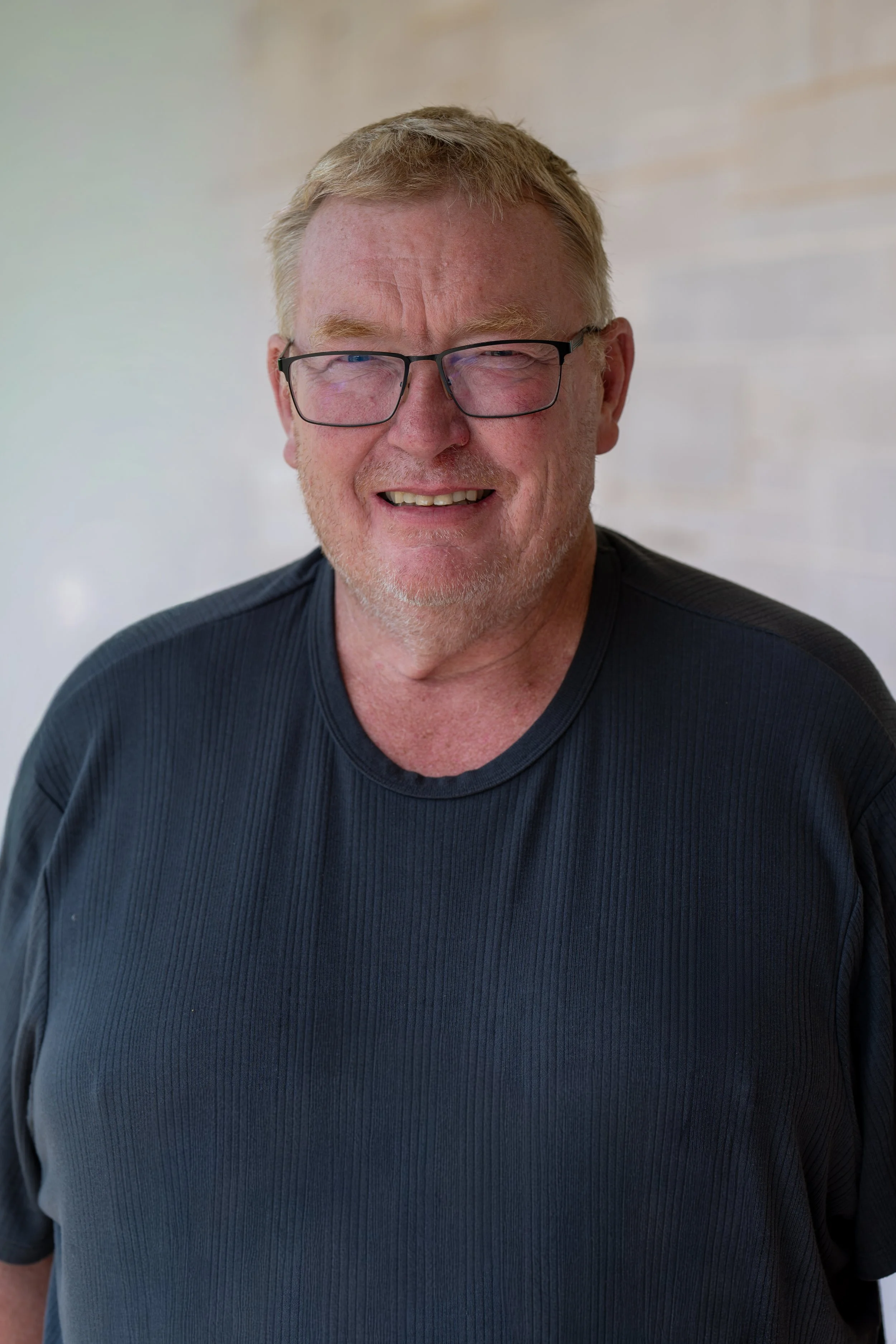 A smiling man with glasses and short hair, wearing a gray shirt with black detailing, posing against a gray background.