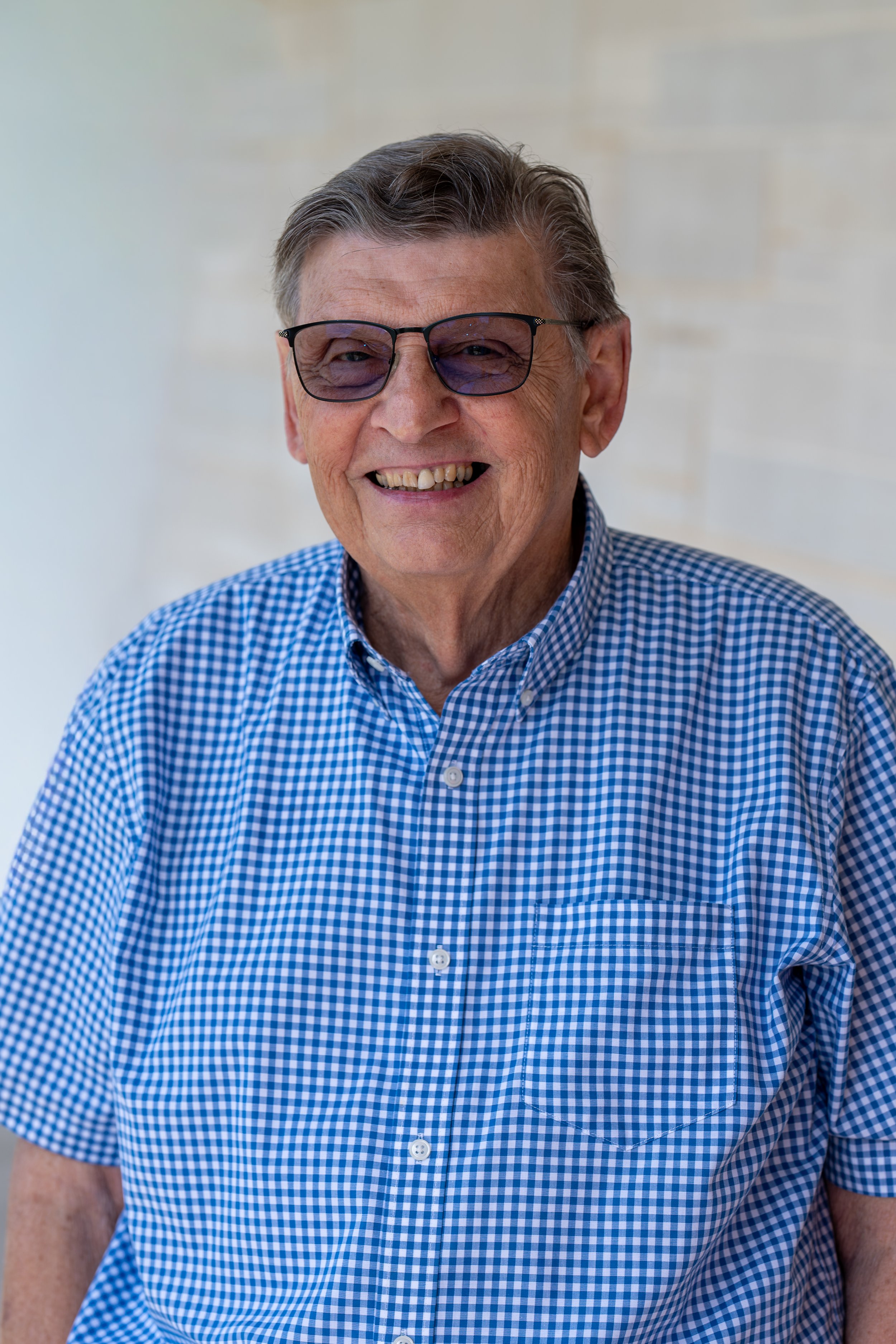 Portrait of a smiling older man with glasses wearing a black polo shirt, against a dark gray background.