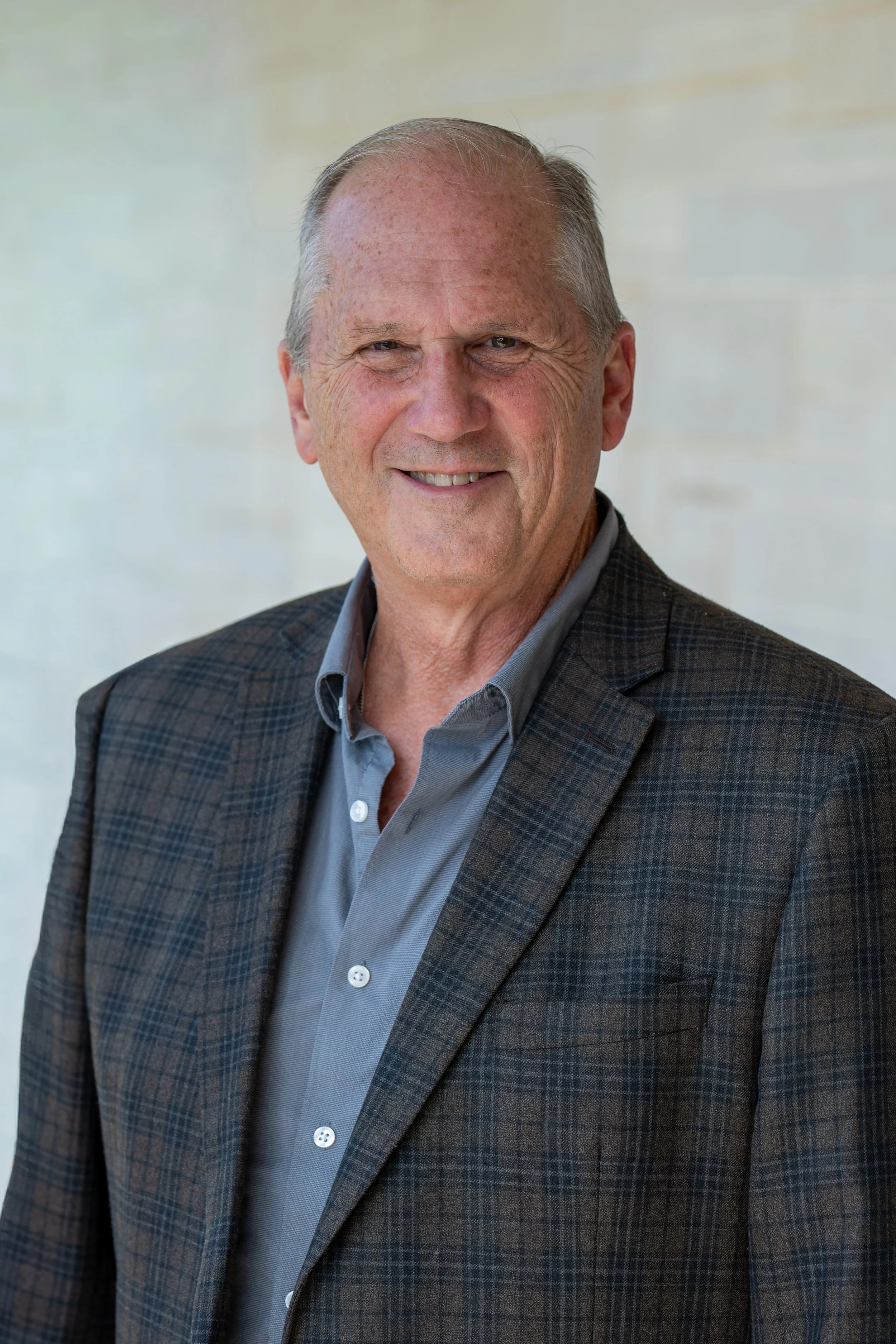 A smiling older man with short, light-colored hair, wearing a beige button-up shirt with a patch on the chest, standing indoors against a brick wall background.