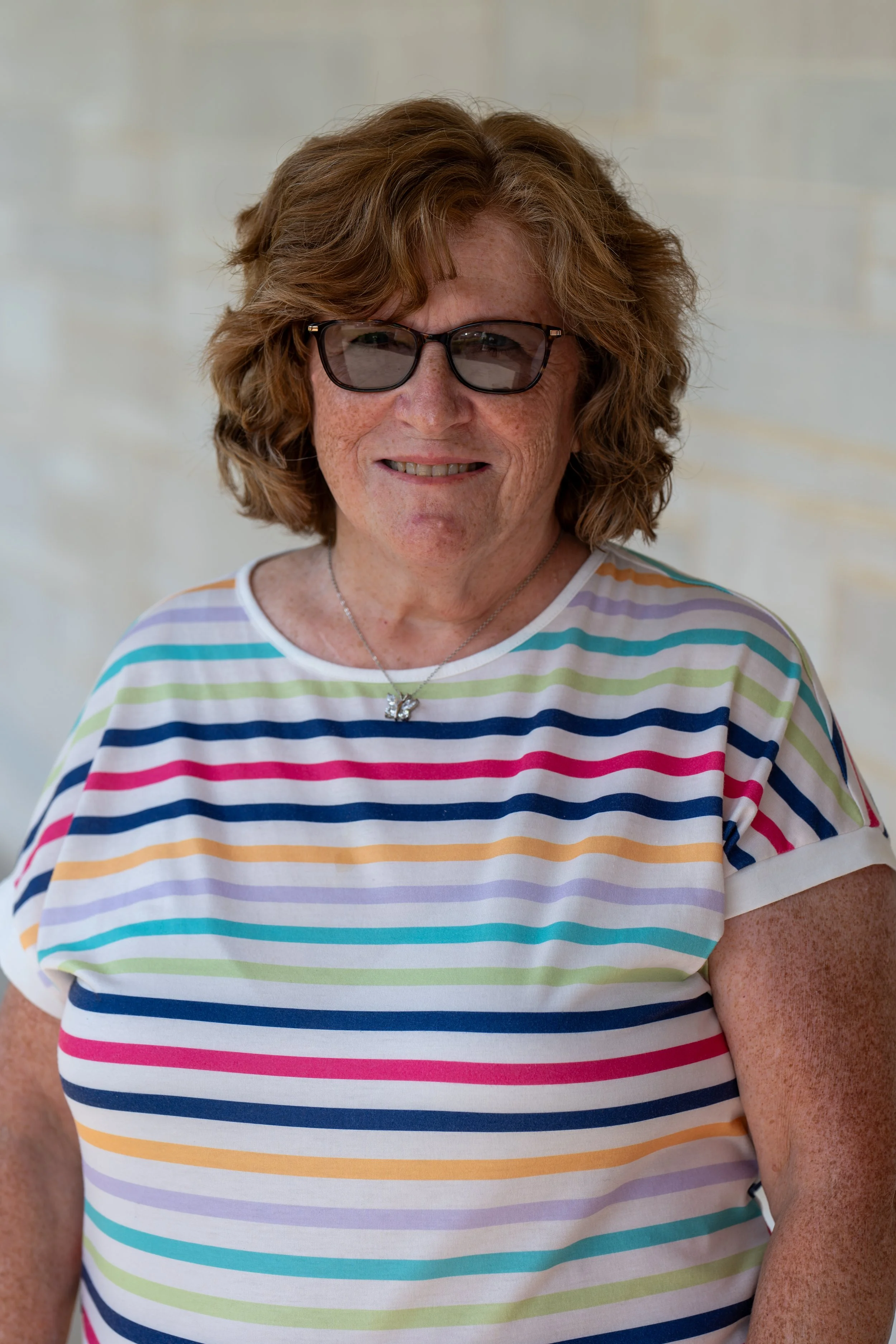 Portrait of a smiling woman with short brown hair, glasses, wearing a blue cardigan and a light-colored top, in front of a gray background.