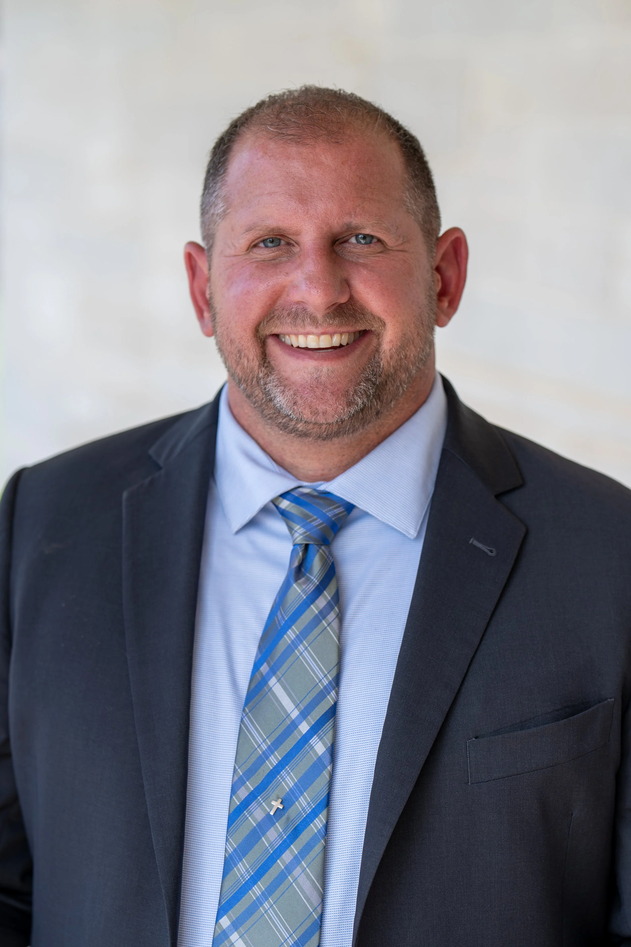 A smiling man with short hair and a beard, wearing a blue button-up shirt, standing in front of a wooden background.