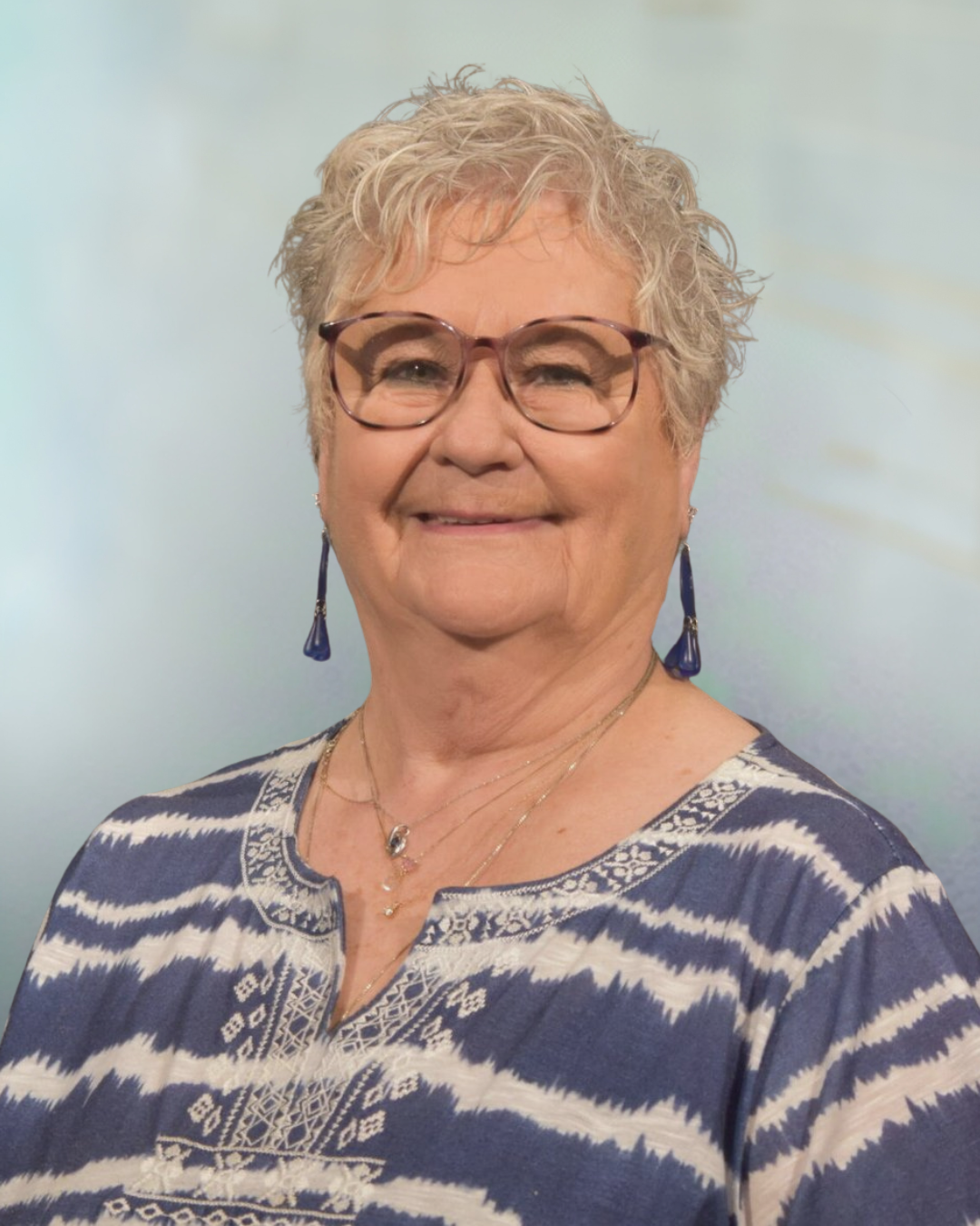 Close-up of a smiling woman with short curly gray hair and glasses, wearing a blue and white patterned top and blue earrings, standing in front of a wooden wall.