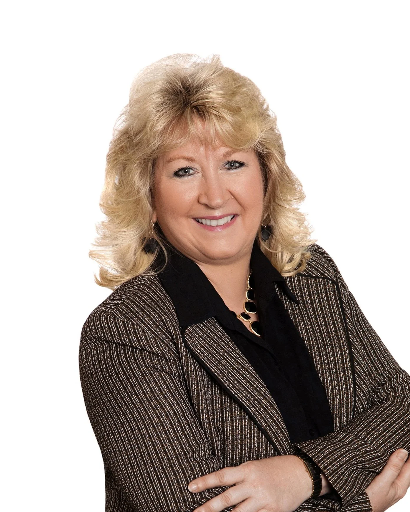 A middle-aged woman with blonde, styled hair, smiling, wearing a black shirt, a gold necklace, earrings, and a brown patterned blazer, posing with her arms crossed against a plain white background.