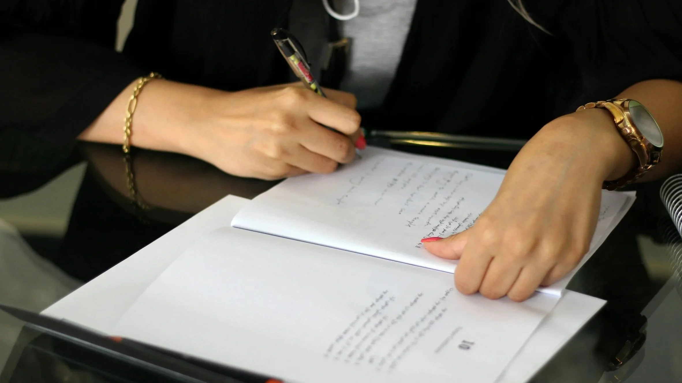 A person writing on a notepad with a pen, resting on a glass table, wearing a gold bracelet and a large watch.