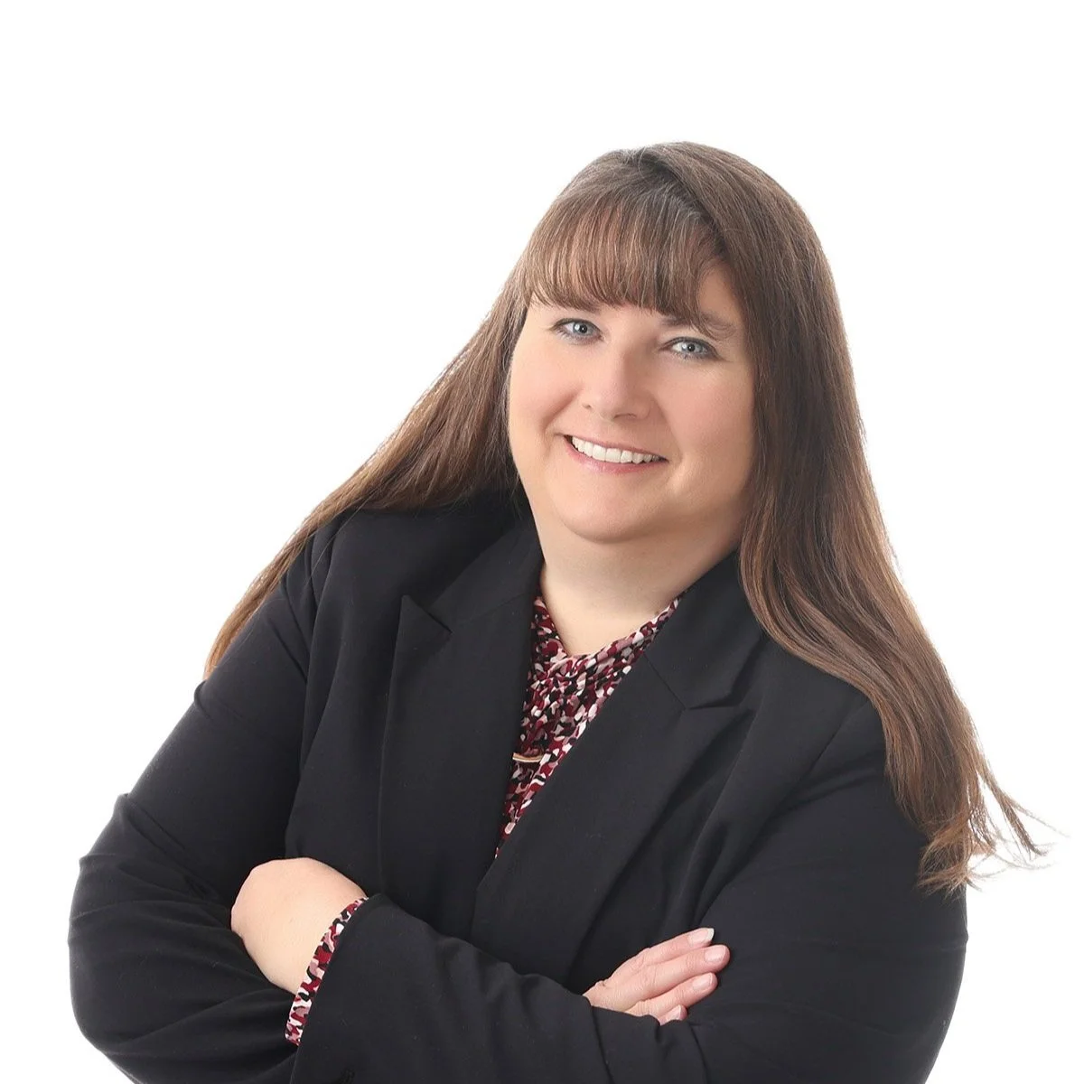 A woman with long brown hair and bangs smiling, wearing a black blazer and patterned blouse, posed with arms crossed against a white background.