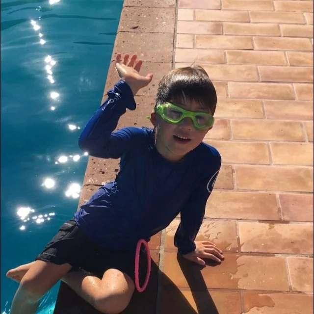 Child in swimwear sitting by a pool waving and wearing green swim goggles