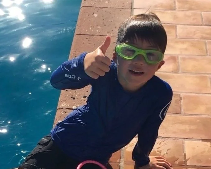 Child in blue rash guard with green goggles giving thumbs up by a pool