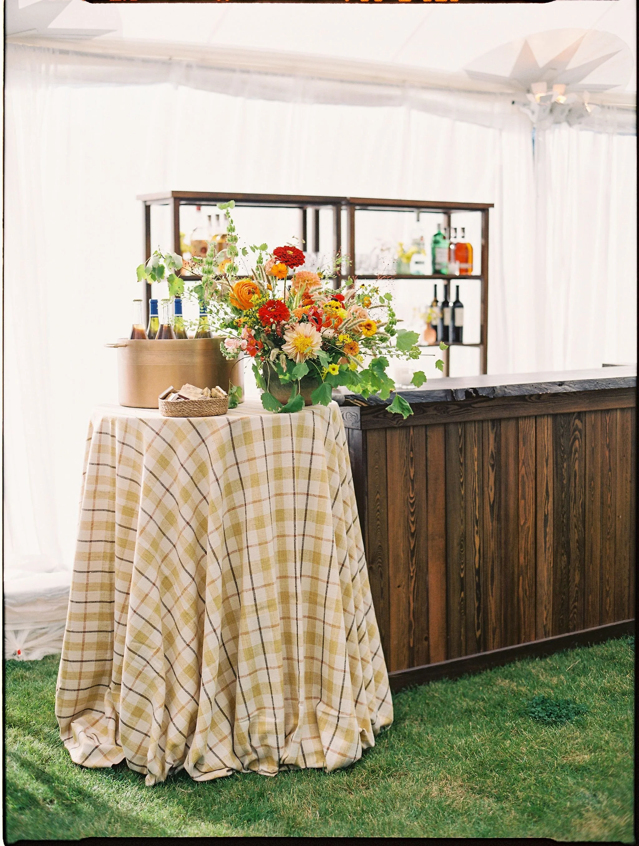 A floral arrangement on a checkered tablecloth with bottles and a basket of napkins, set next to a wooden bar inside a tent.