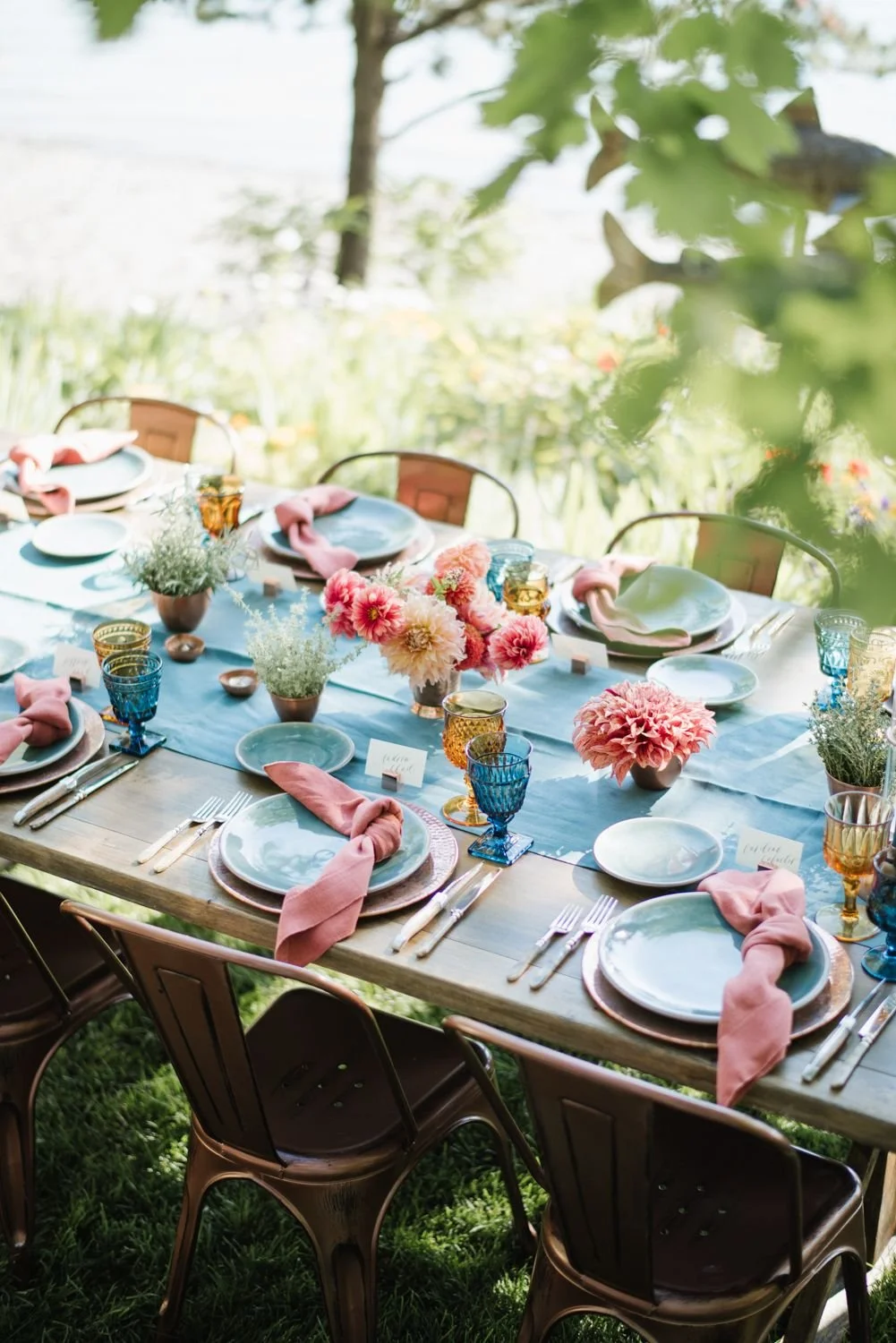 An outdoor dining table decorated with pink and peach flowers, pink napkins, and colorful glassware, set for a gathering.