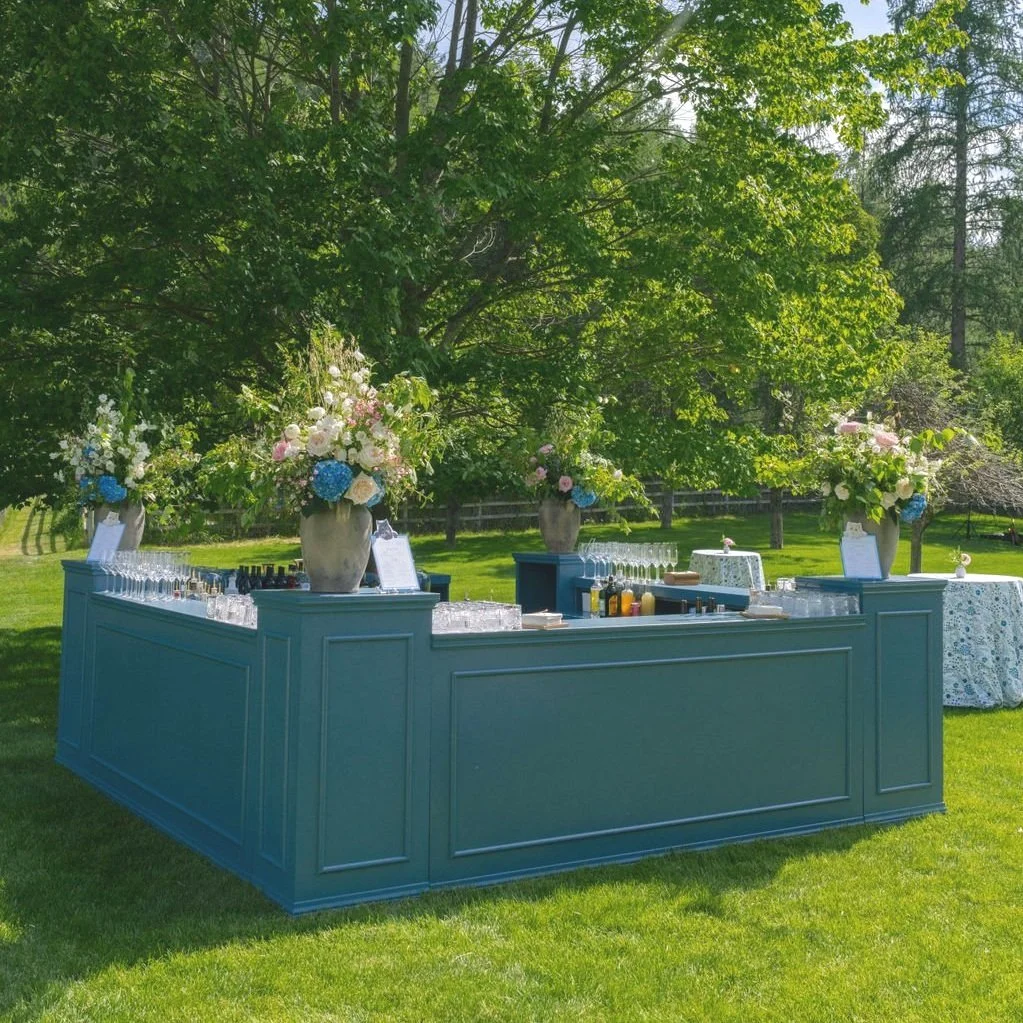 An outdoor bar with floral arrangements, glasses, and bottles set up under a large green tree on a sunny day.