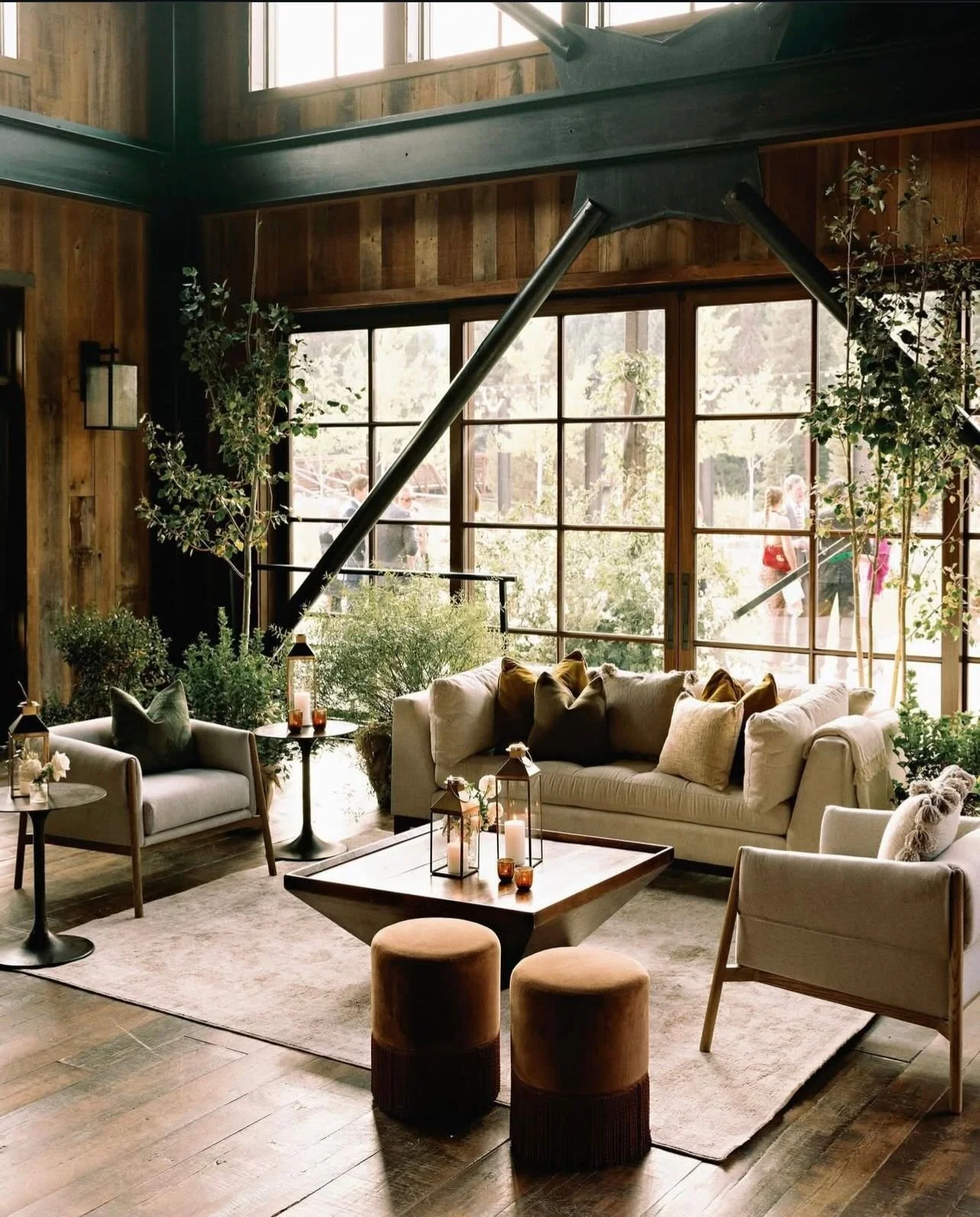 A cozy living room with beige sofas, armchairs, and a wooden coffee table in front of large windows, decorated with candles and lanterns, with plants and wooden walls.