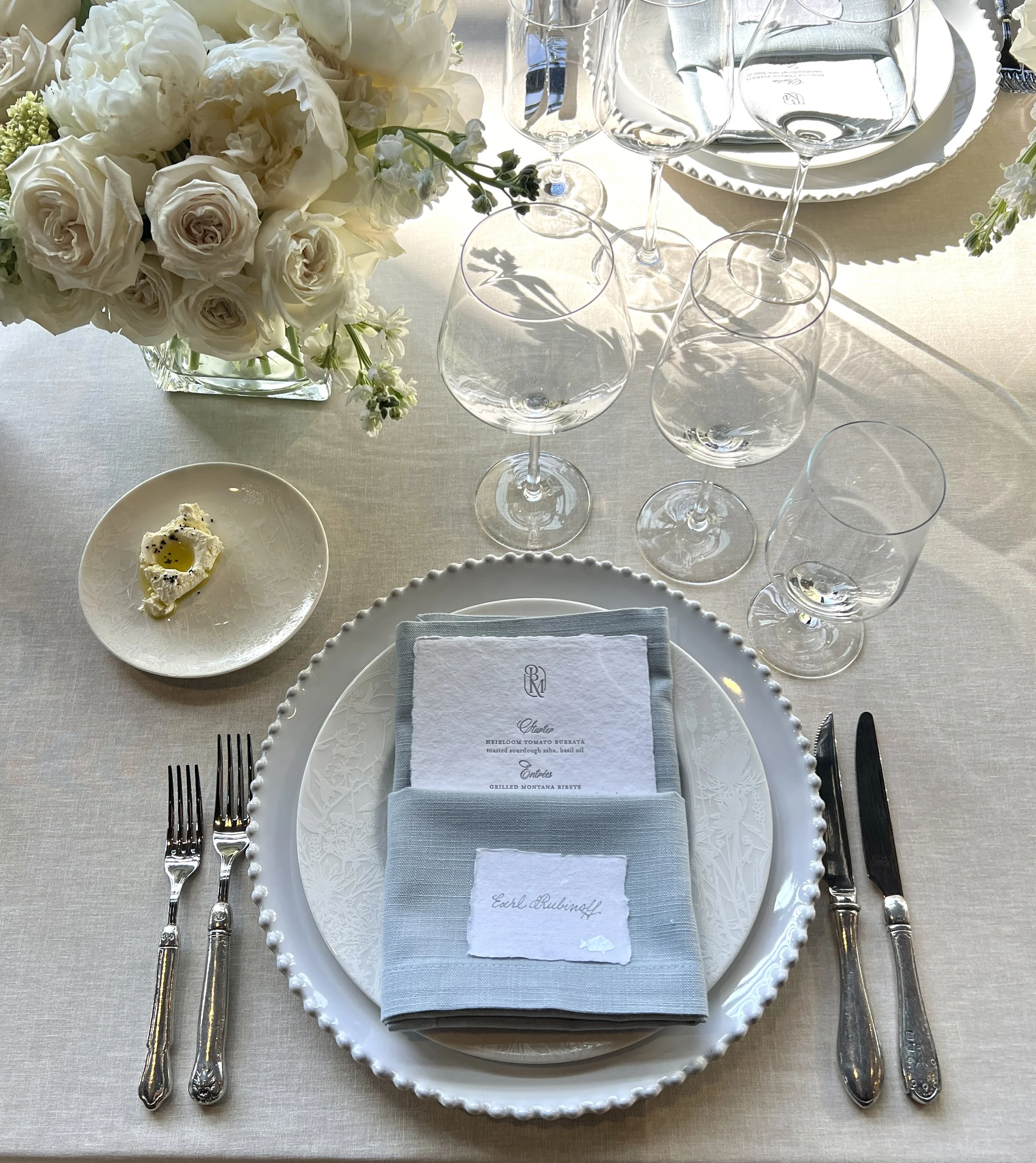 Elegant table setting with a white floral centerpiece, clear glassware, silver cutlery, a decorative white plate, a grey napkin with a place card, and a small dish with butter and olive oil, set on a white tablecloth.