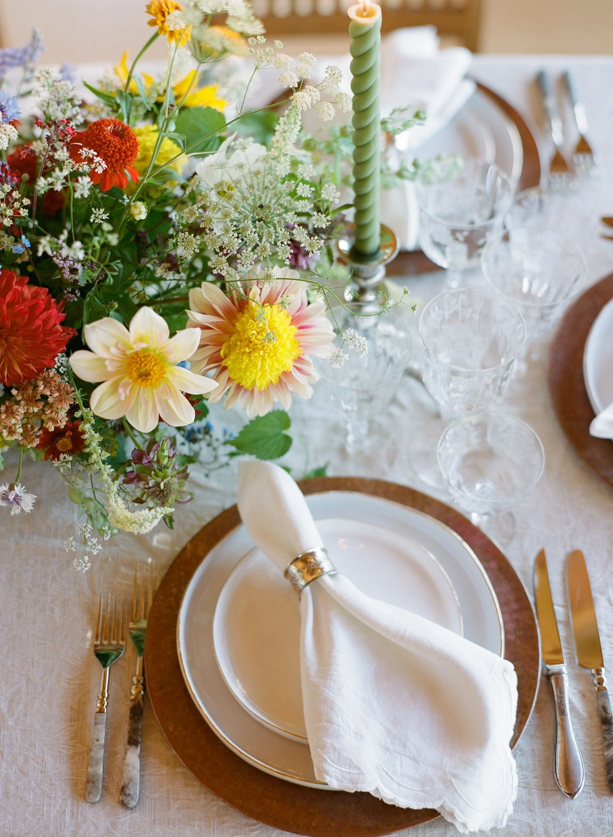 Elegant dining table with a floral centerpiece, white plates with gold trim, a white napkin with a silver napkin ring, crystal glasses, and silverware, with a tall green twisted candle.