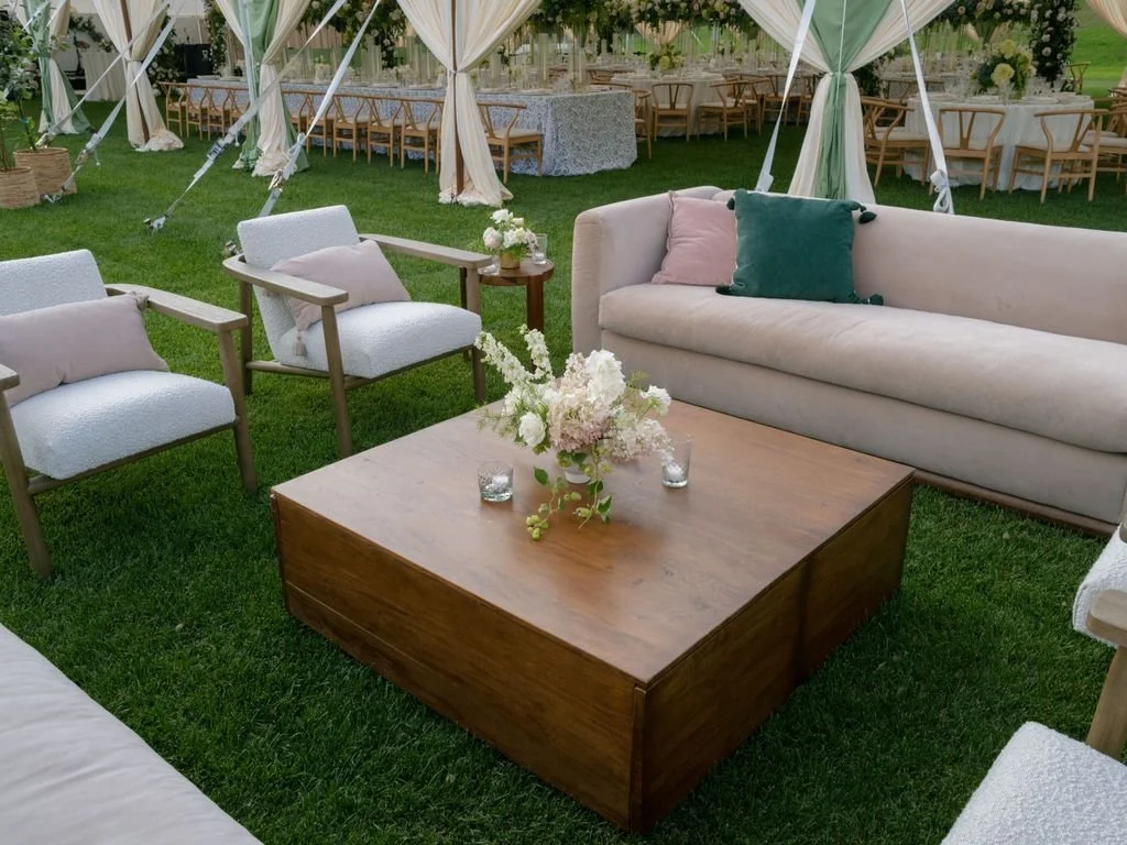Outdoor event setup with beige sofa, white chairs with beige pillows, wooden coffee table with floral centerpiece and candles, tents with draped fabric, tables with floral centerpieces and chairs in the background.