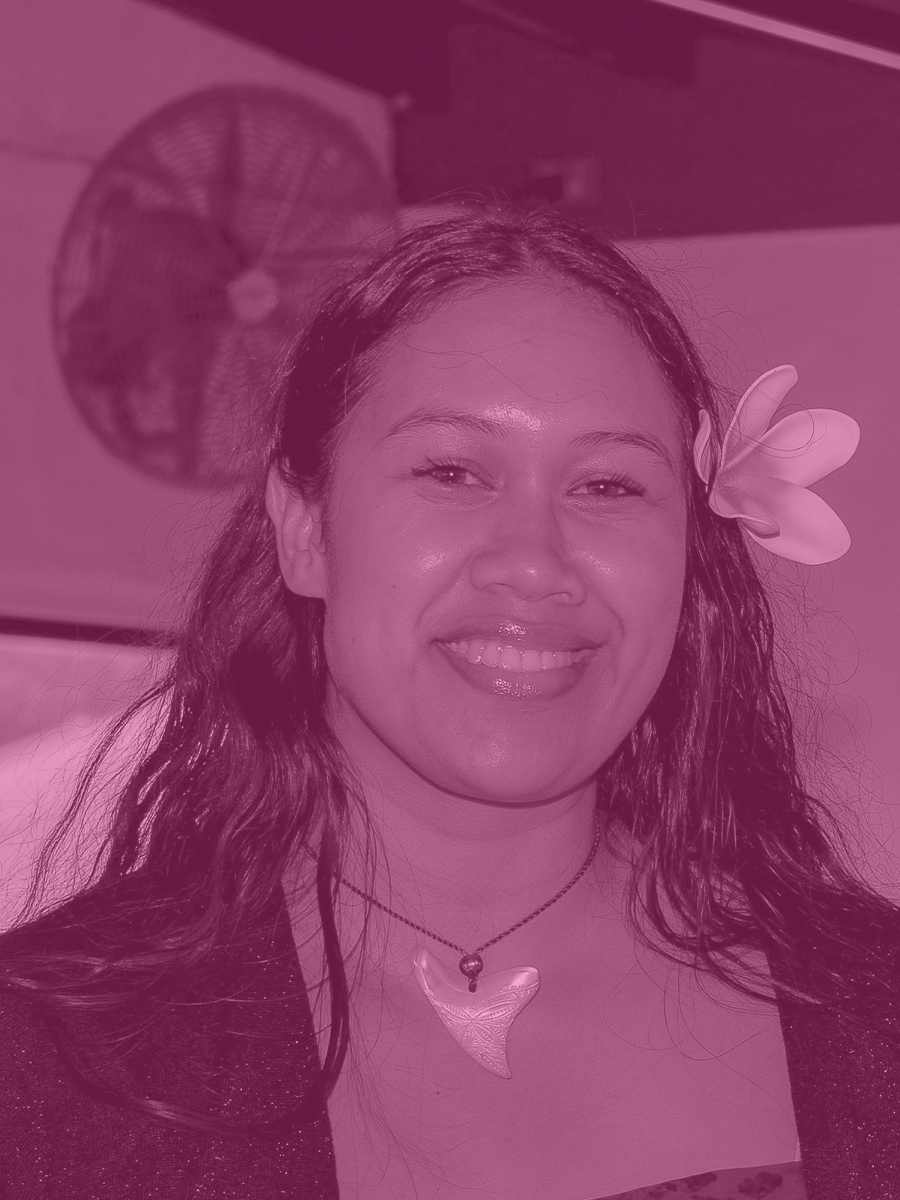 A woman with long hair and a large flower in her hair, smiling, wearing a necklace with a shell pendant, indoors against a wall with a fan in the background.