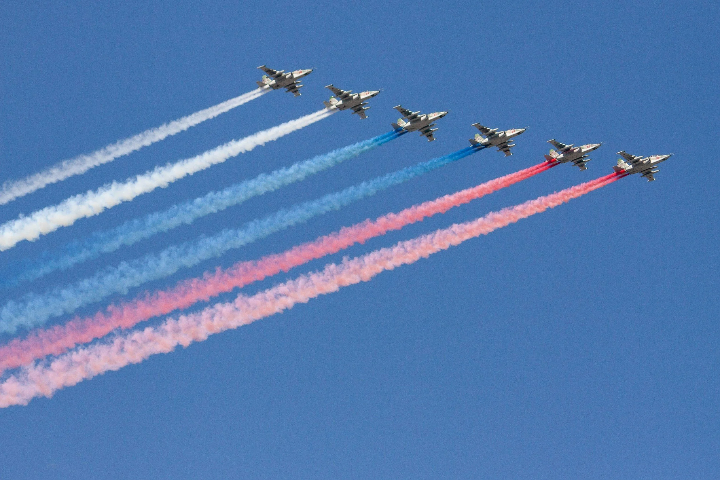 Six fighter jets flying in formation with red, blue, and white smoke trails against a clear blue sky.