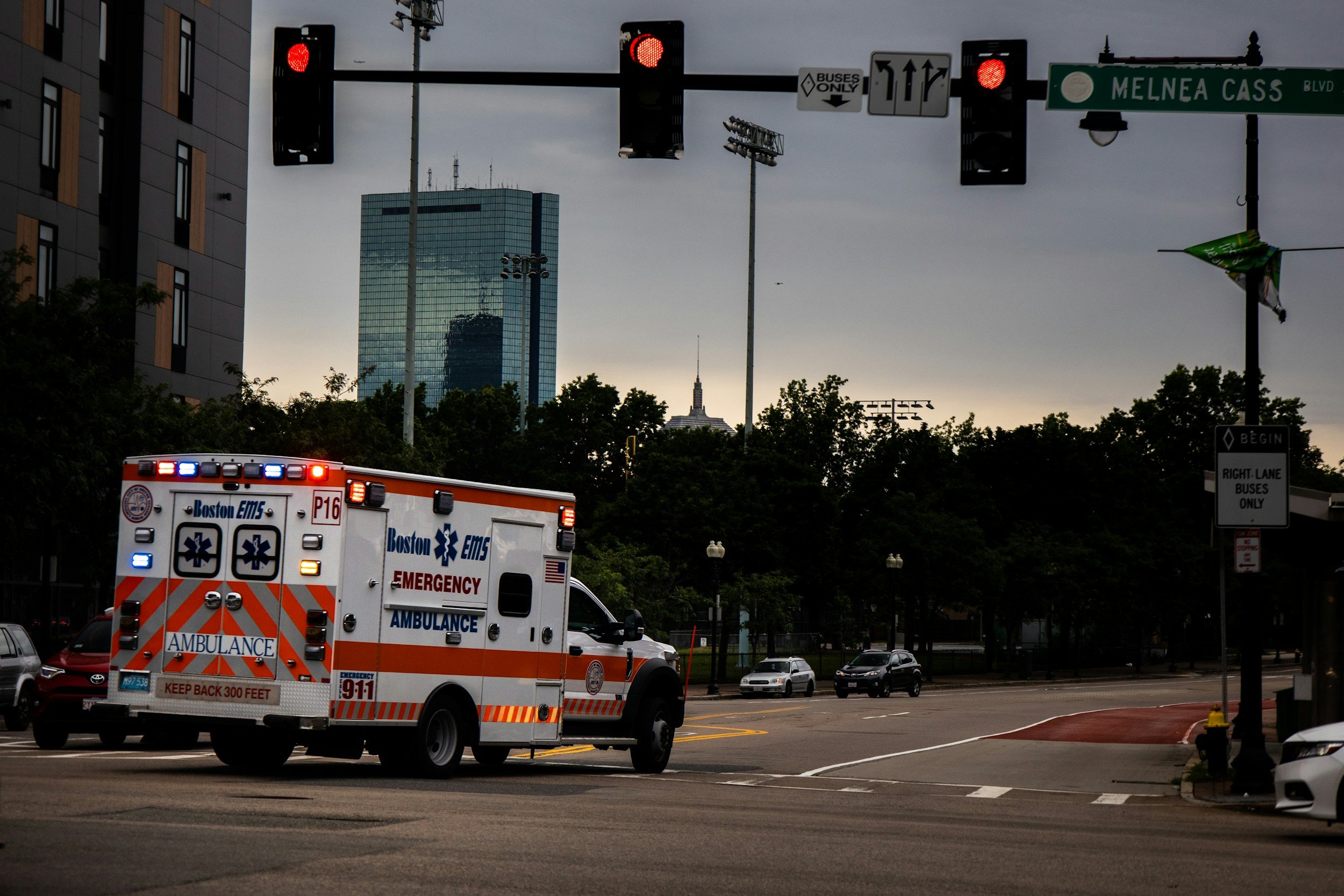 Boston EMS ambulance stopped at a red light on Melnea Cass Boulevard, with city skyline and trees in the background.