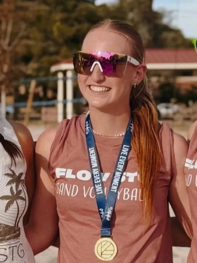 A young woman with red hair tied back in a ponytail, smiling, wearing a black t-shirt with a circular logo that says "Flow STG EST 2025," standing outdoors near a complex structure with glass and several poles.