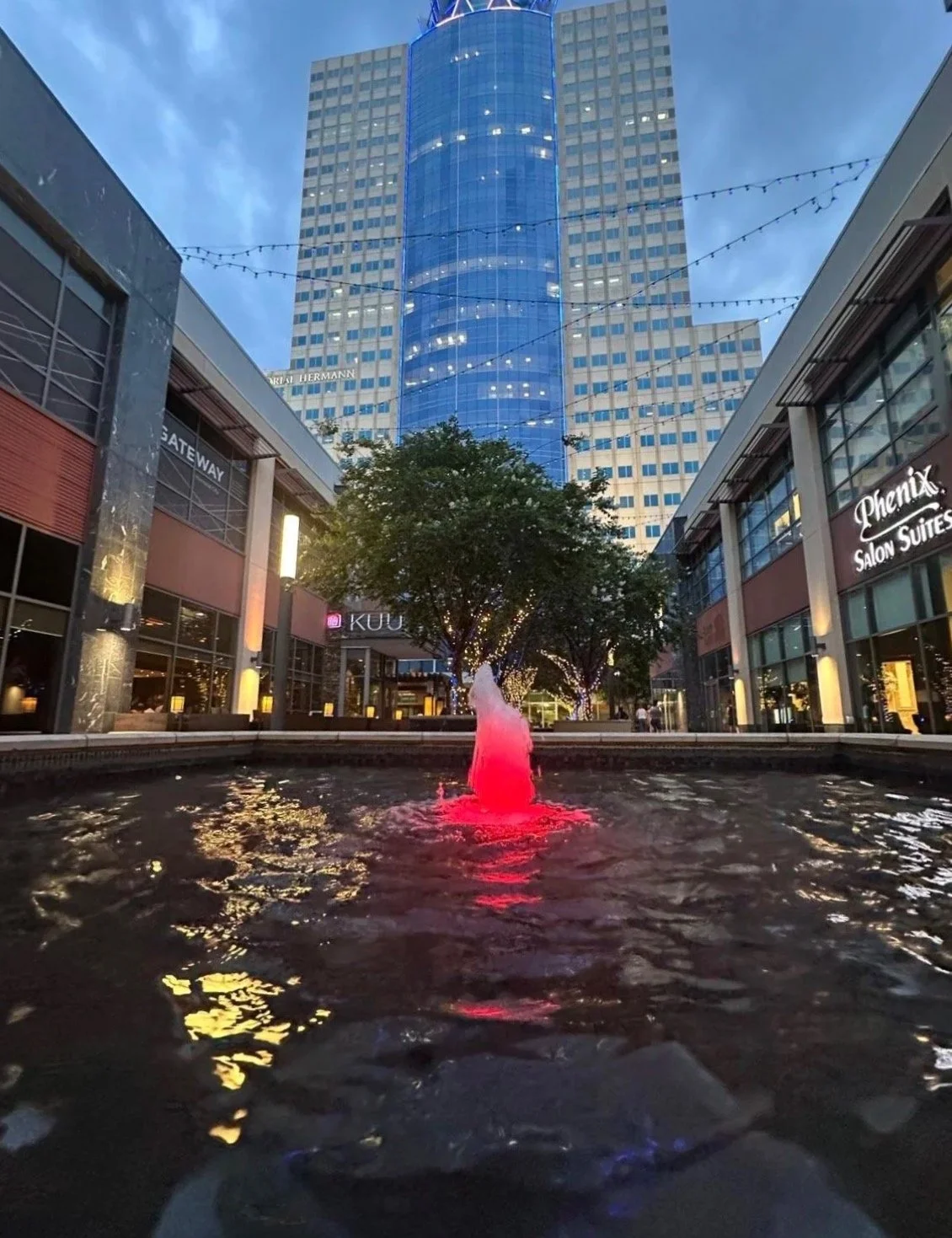 A modern outdoor shopping mall with a central fountain illuminated with pink light, surrounded by trees decorated with string lights, and tall buildings in the background.