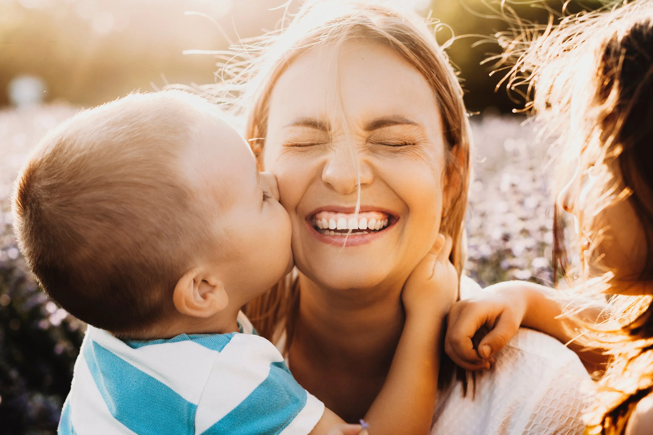 A woman smiling with eyes closed being kissed on the cheek by a young boy with blonde hair, wearing a blue and white striped shirt, outdoors during sunset.
