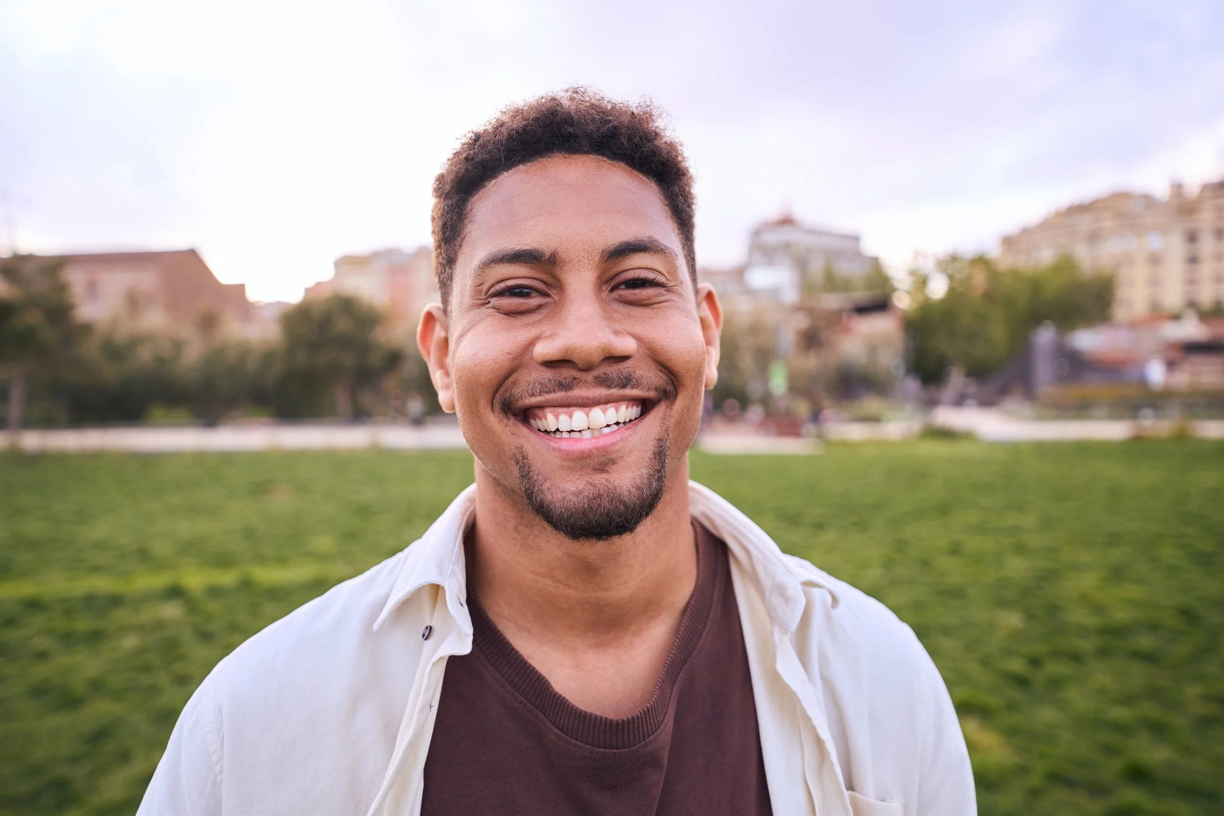 Smiling man standing in a park with buildings in the background under a cloudy sky.