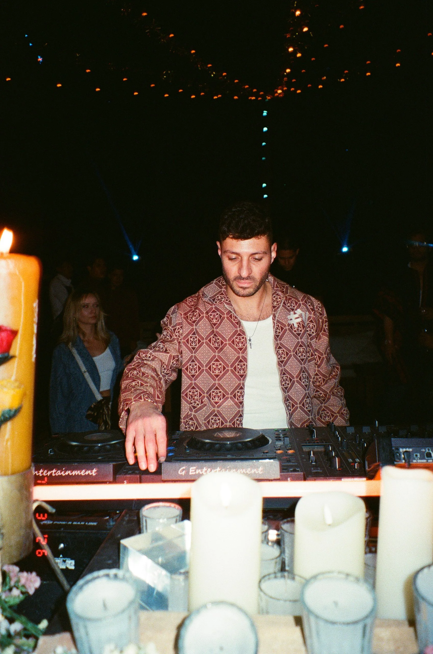 DJ Niccc, with short curly hair and a patterned shirt is playing music at Amarás Oaxaca Experience 2026, with candles, flowers, and people dancing in the background.