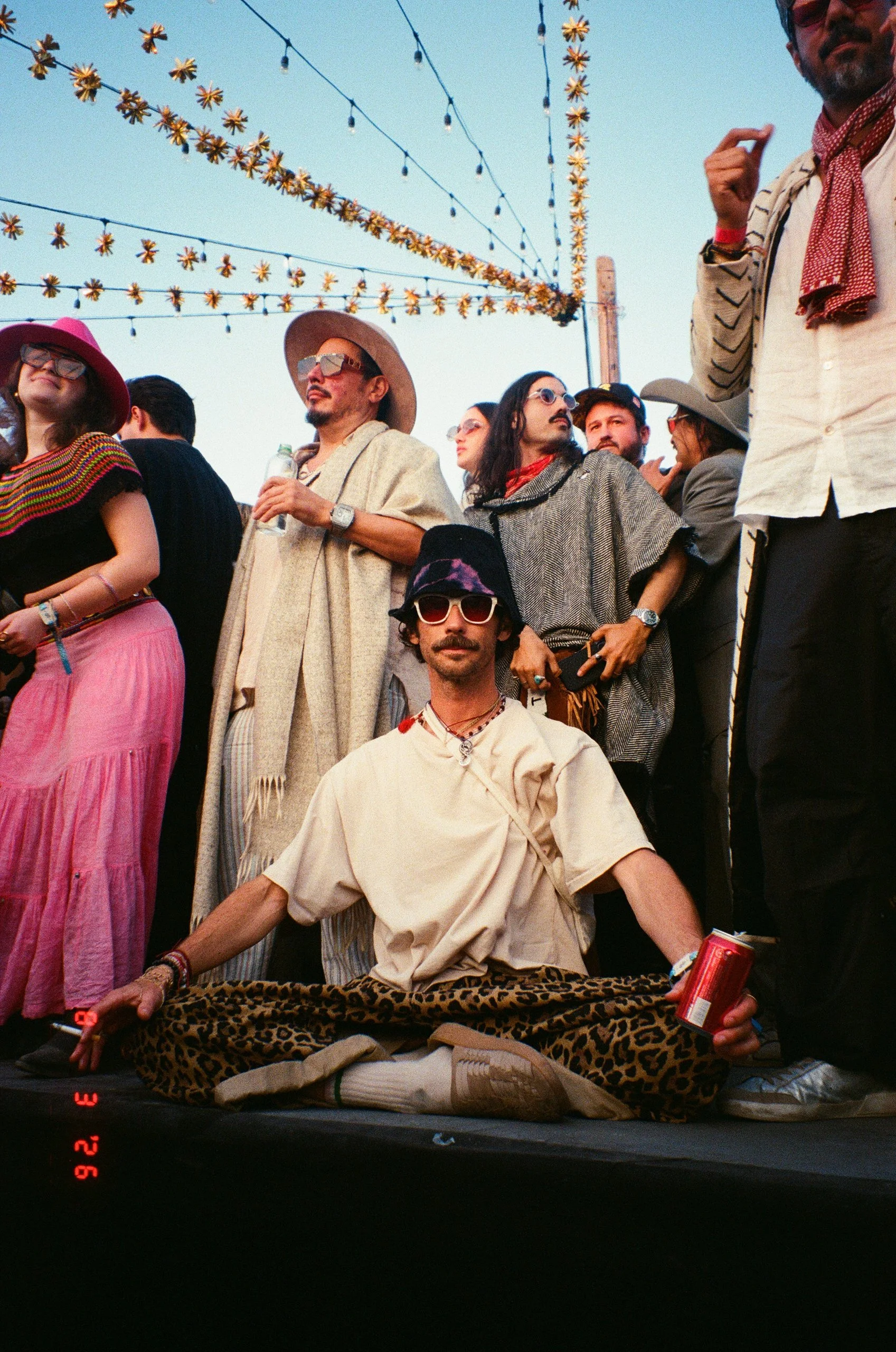 A group of people with diverse styles and accessories at Amarás Oaxaca Experience 2026, some standing and one person sitting cross-legged on a leopard-print blanket, holding a drink, with string lights above.
