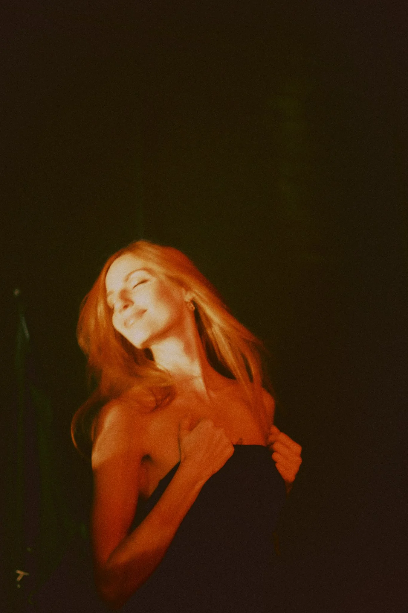 A woman with long red hair smiling and closing her eyes, wearing a black strapless top, holding it with both hands against a dark background.