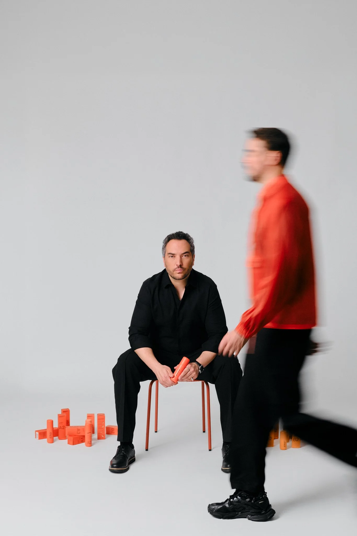 Studio portrait shoot featuring two men against a seamless white background. One man sits centered on an orange metal stool, dressed in all black, holding an orange cylindrical product and looking directly into camera with a calm, composed expression