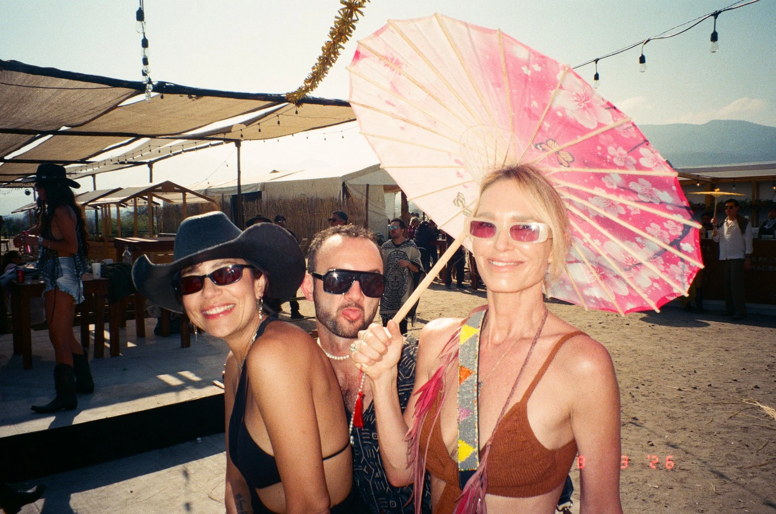 Three smiling people at an outdoor event, with two women wearing sunglasses and one with a pink floral umbrella. Background shows outdoor seating and other people at Amarás Oaxaca Experience 2026.
