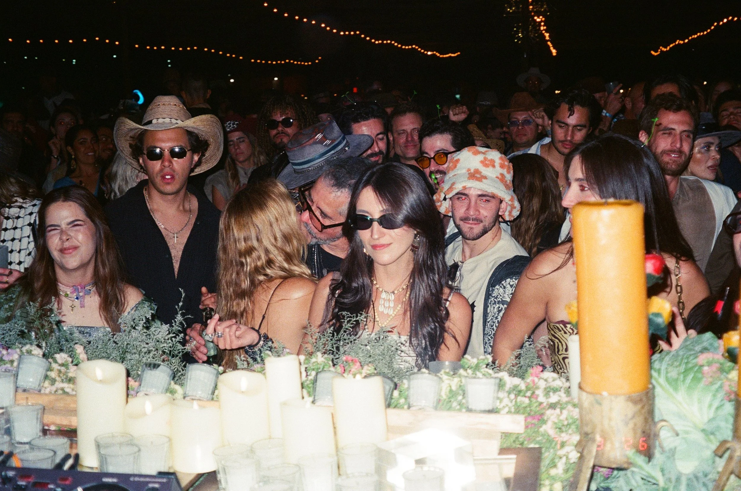A crowd of people gathered at Amarás Oaxaca Experience 2026, with candles and floral decorations in the foreground, illuminated by string lights overhead, some wearing sunglasses and hats.