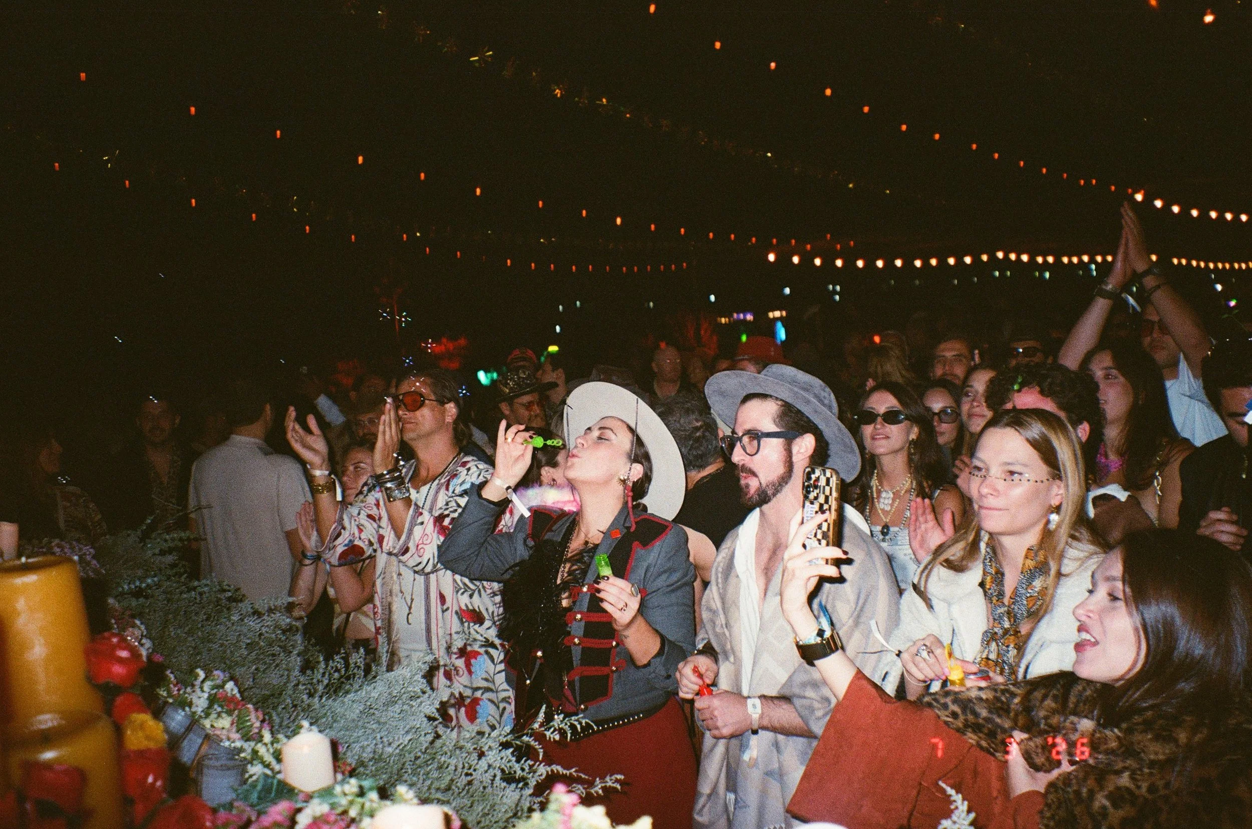 A crowd of people at Amarás Oaxaca Experience 2026, dressed in colorful clothing, with some wearing hats and sunglasses, under string lights at night.
