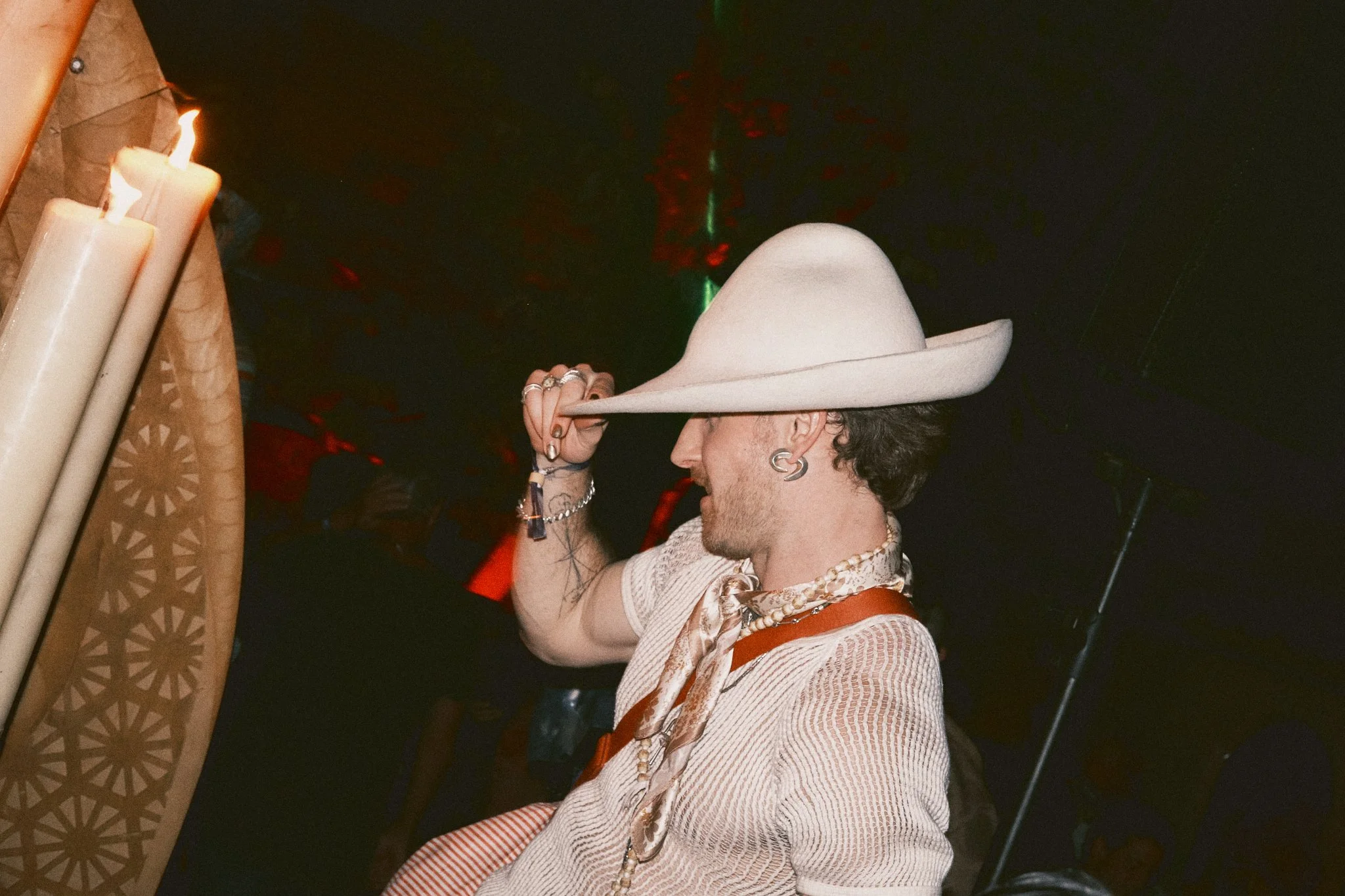 A person wearing a large white hat, earrings, and layered necklaces, tipping the hat in a dimly lit setting with candles on the side.
