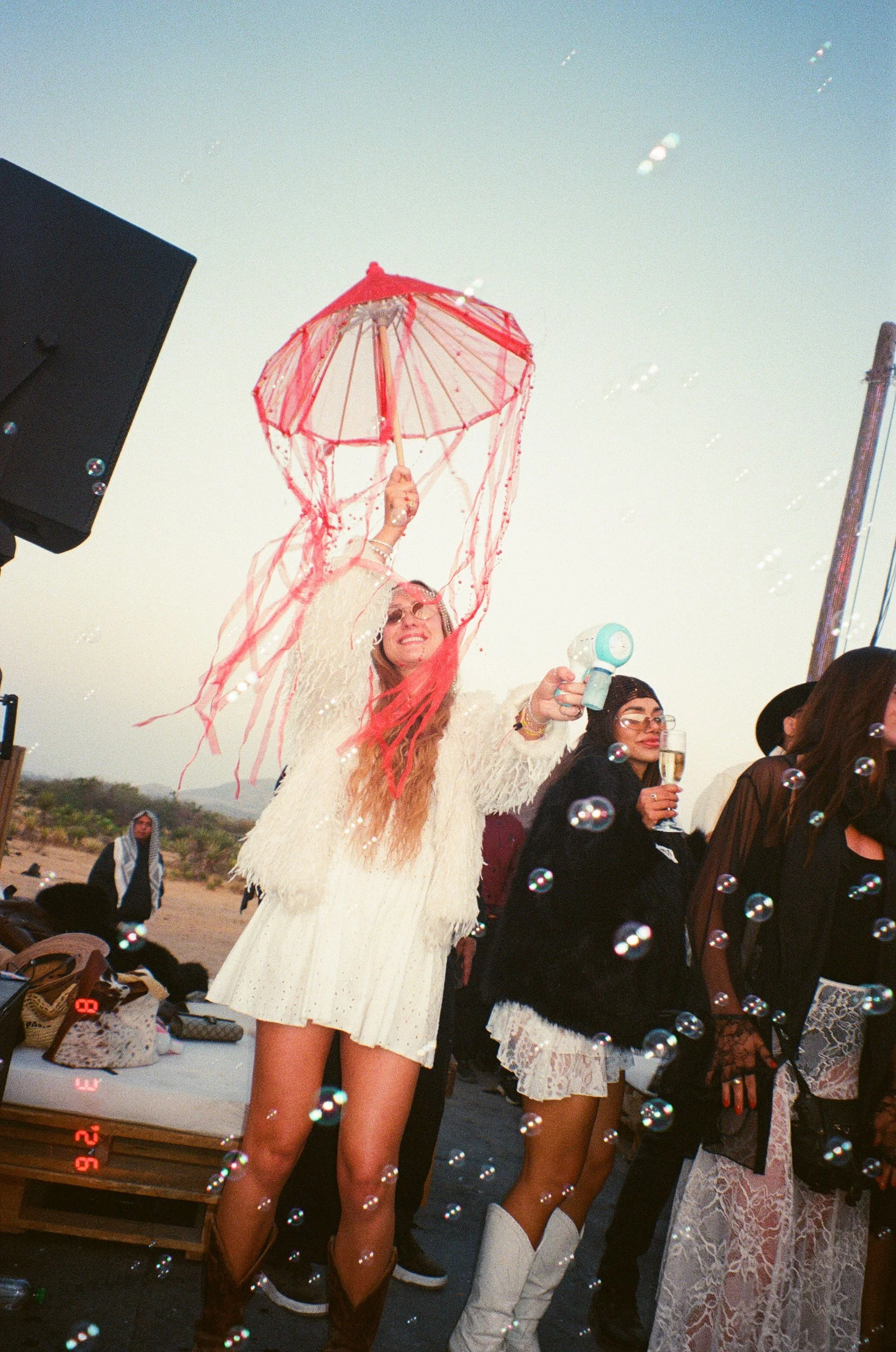 Group of women at Amarás Oaxaca Experience 2026, one woman in the foreground holding a red parasol and a handheld fan, others holding glasses with drinks, surrounded by bubbles, with the sky in the background.