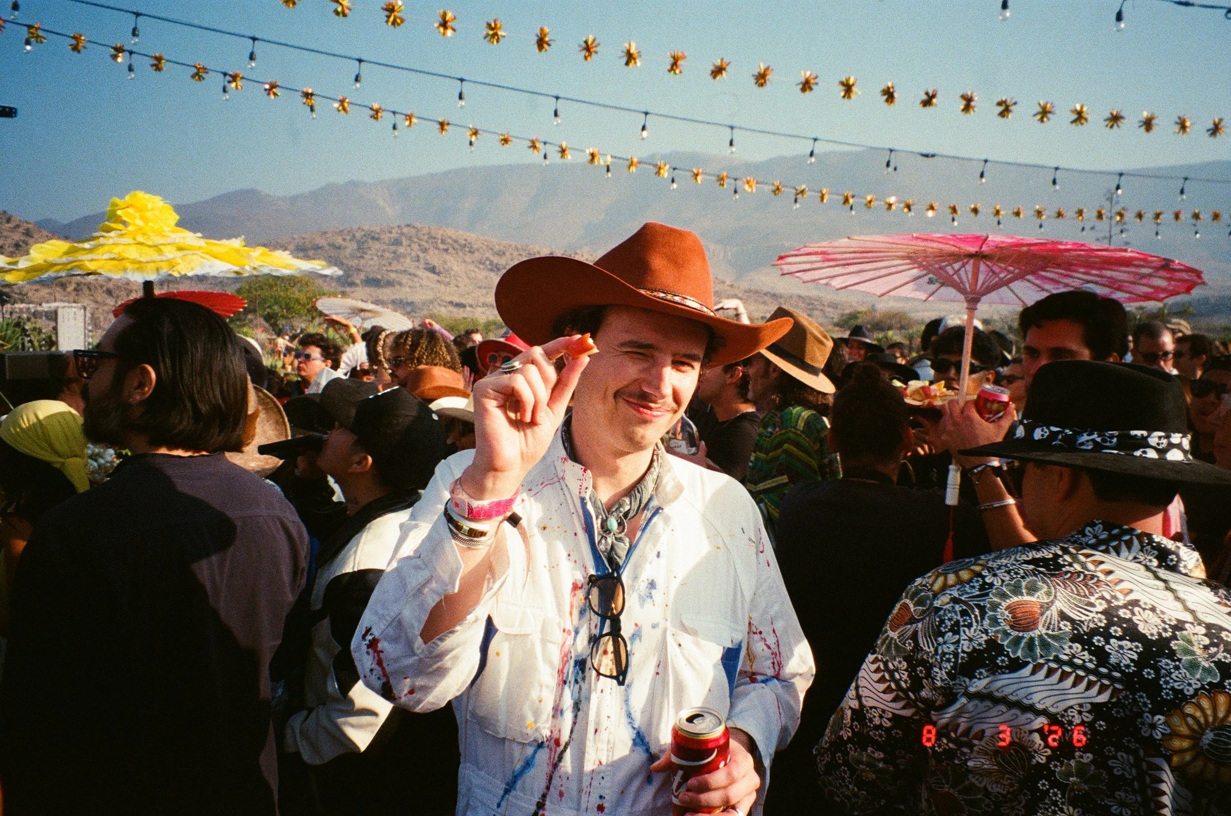Salimbeni in the center is wearing a cowboy hat, smiling, and holding an apple slice. People in the background are wearing hats and sunglasses, and there are string lights and colorful umbrellas overhead at Amarás Oaxaca Experience 2026.