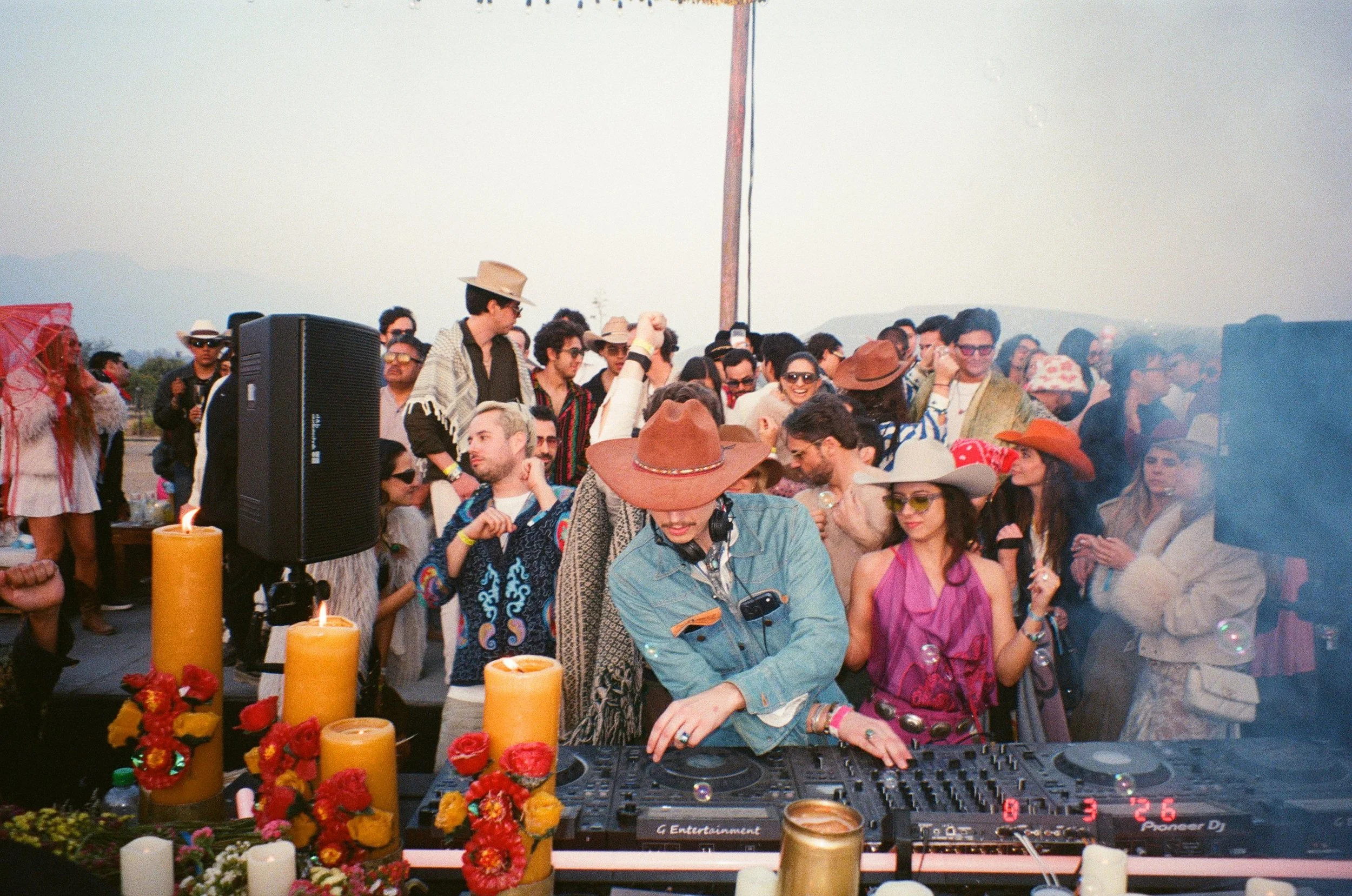 DJ Salimbeni at Amarás Oaxaca Experience 2026. A diverse group of people behind him and candles and flowers, on the booth during the daytime.