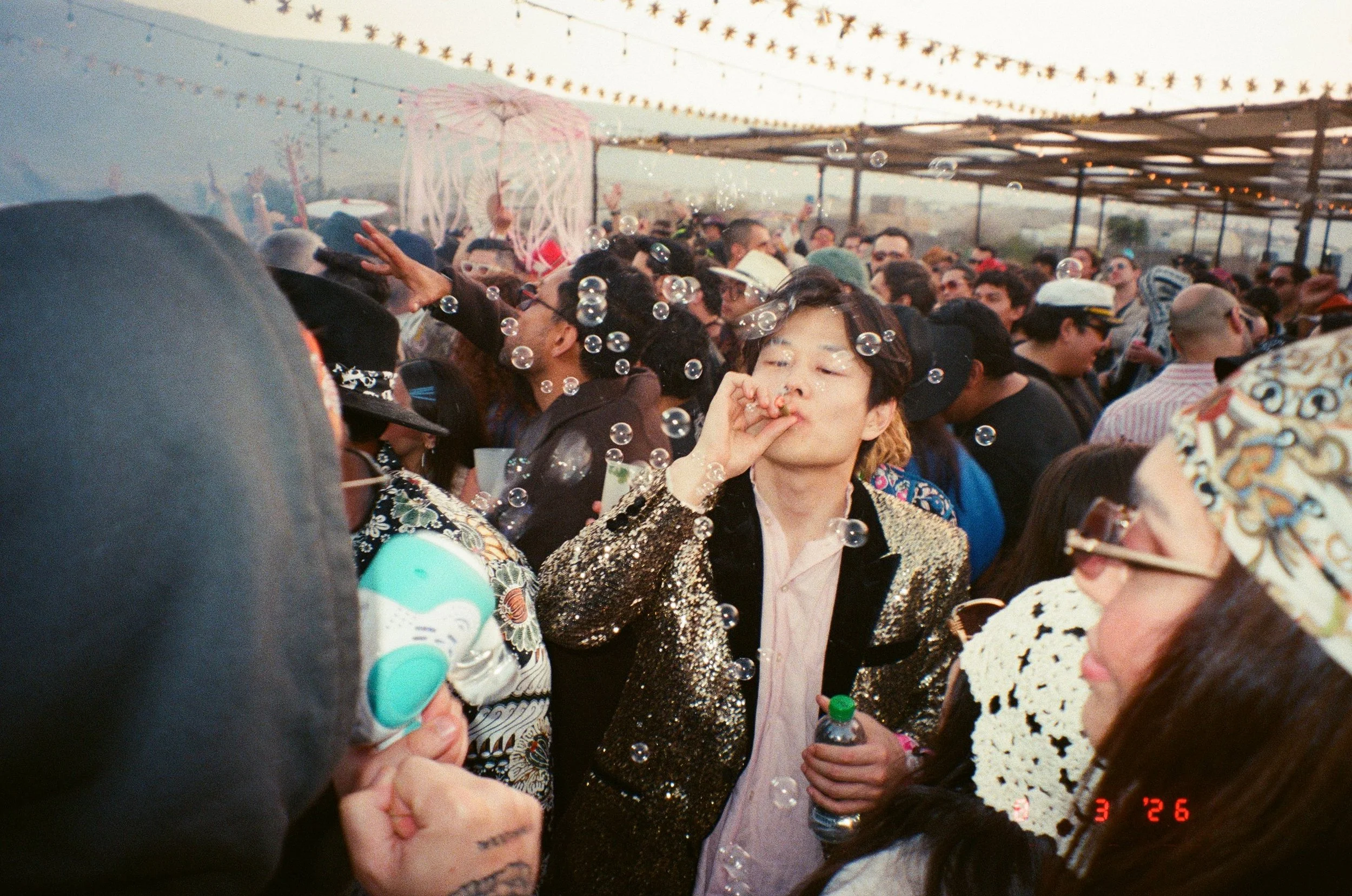 A large crowd of people celebrating at Amarás Oaxaca Experience 2026, some blowing bubbles and wearing colorful and patterned clothing, under string lights during sunrise.