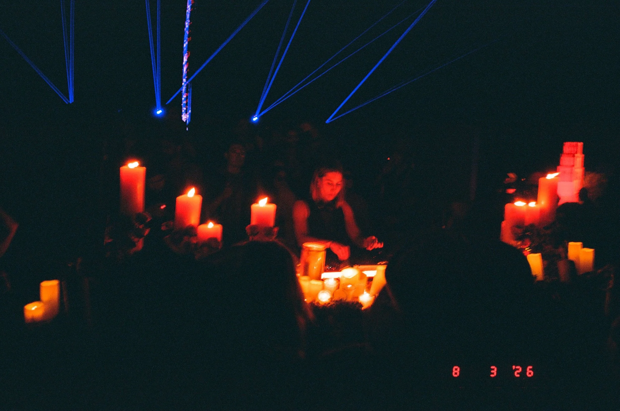 A dark DJ booth at night with several lit candles on a table, LENI playing at Amarás Oaxaca Experience 2026, people standing around, and blue laser lights projecting patterns in the air.