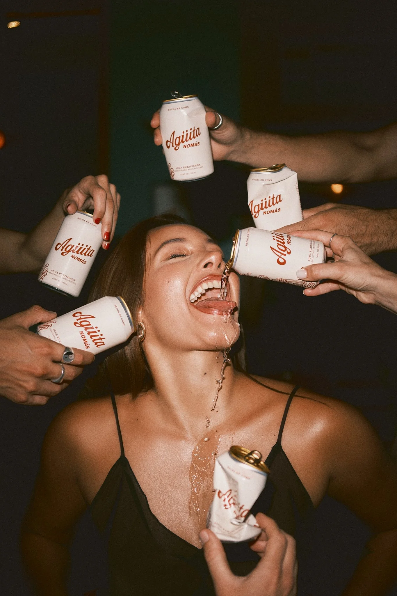 Editorial campaign shot of a woman joyfully tilting her head back with her mouth open as five hands simultaneously pour and hold Agüita Nomás purified water cans around her. Water spills down her face and chest. Dark, warm-toned party atmosphere. Sho