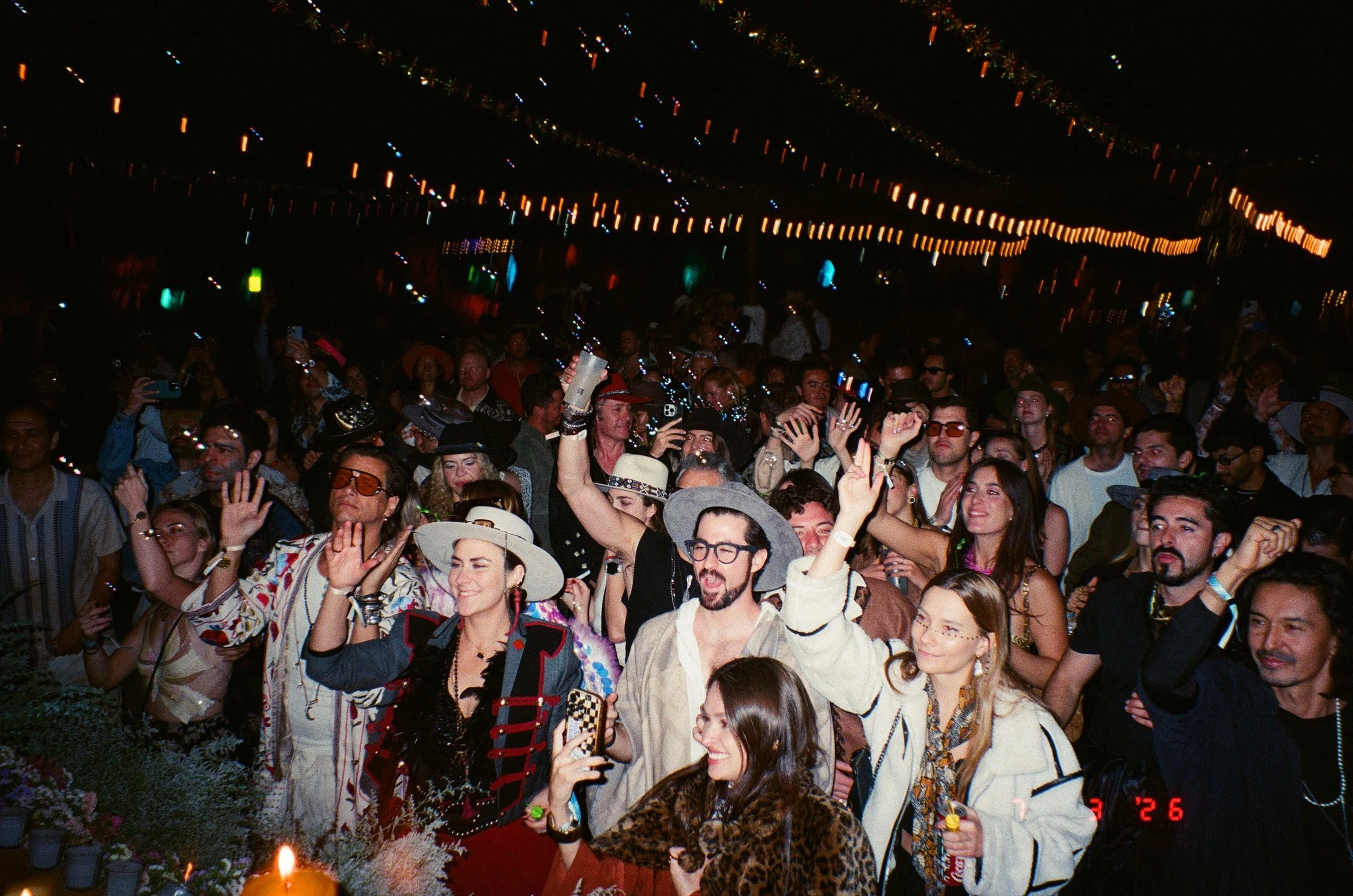 A large crowd of people at Amarás Oaxaca Experience 2026. Some individuals are wearing hats, sunglasses, and colorful clothing. String lights illuminate the scene, and a flower arrangement is visible in the foreground.