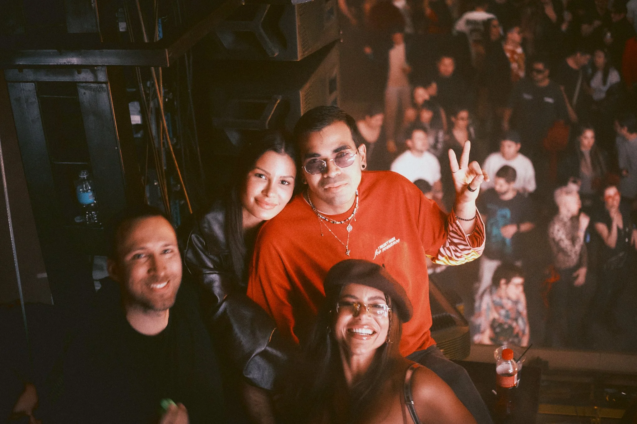 Four people, two men and two women, are posing for a photo backstage at a concert or event. The group includes a man wearing sunglasses making a peace sign, a woman with long dark hair, a man with a bald head, and a woman wearing a beret and glasses.
