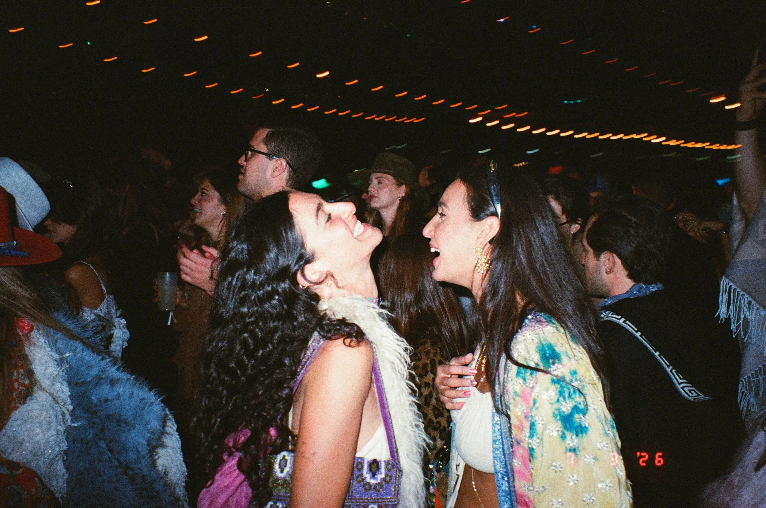 Two girls in the foreground and a group of diverse people in the background, dancing and enjoying live music at Amarás Oaxaca Experience 2026, with string lights overhead and a dark background.