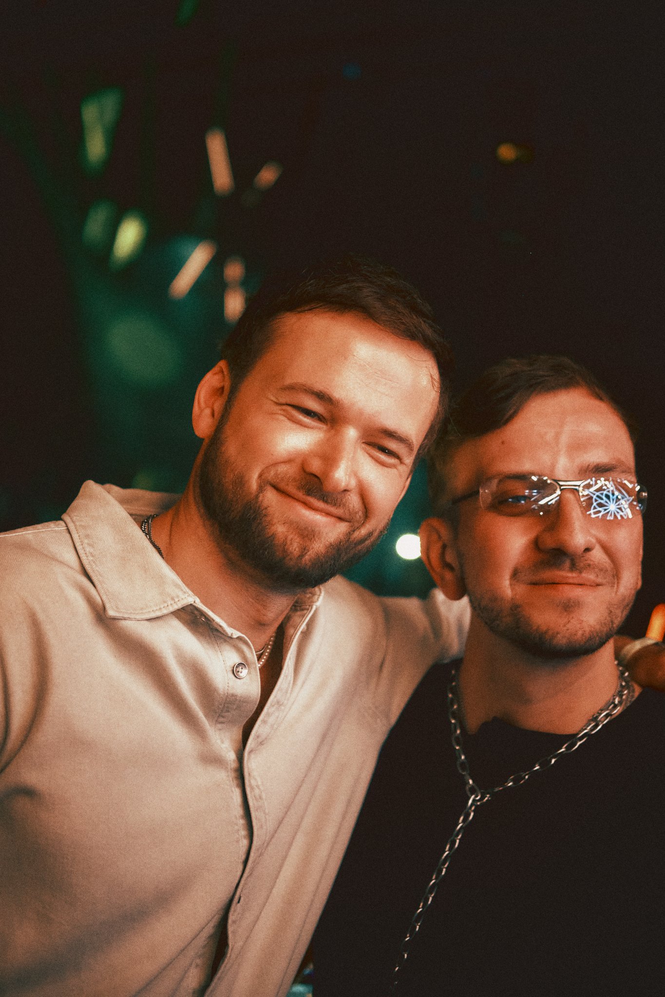 Monolink and Colyn smiling and posing backstage of Mayan Warrior Art Car during Art Week in Mexico City 2026, with one wearing glasses and chains, and the other with a short haircut and beard, illuminated by colorful lights.
