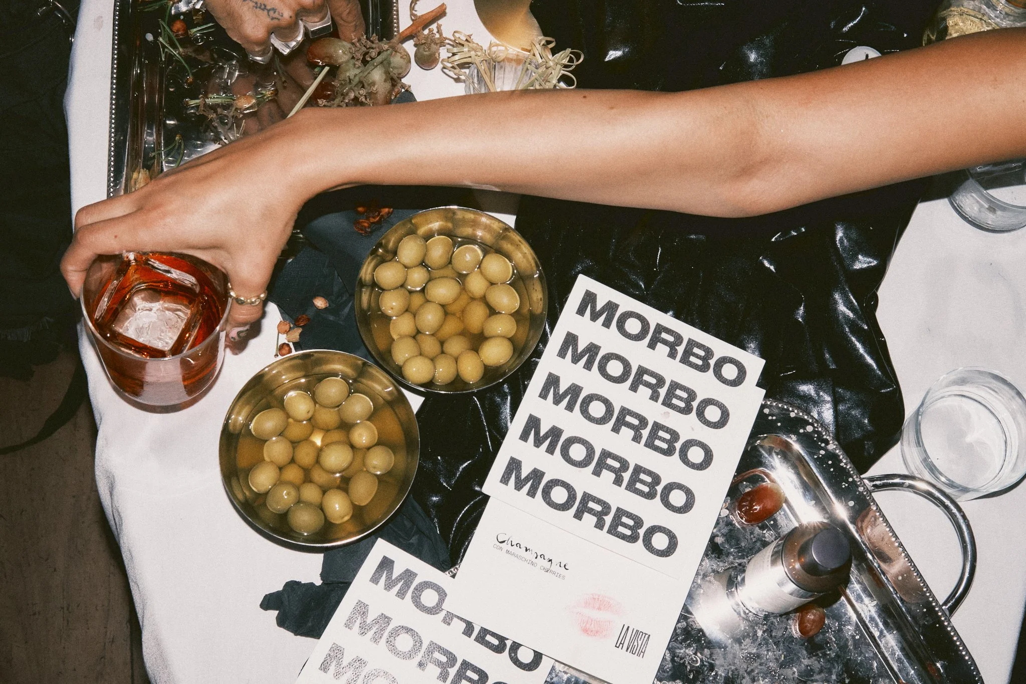 Editorial detail shot from above of a party table at Morbo, an event at La Vista in Mexico City. A hand holds a whiskey on the rocks while two silver bowls filled with green olives sit beside stacked Morbo-branded menus. A champagne bottle chills on 