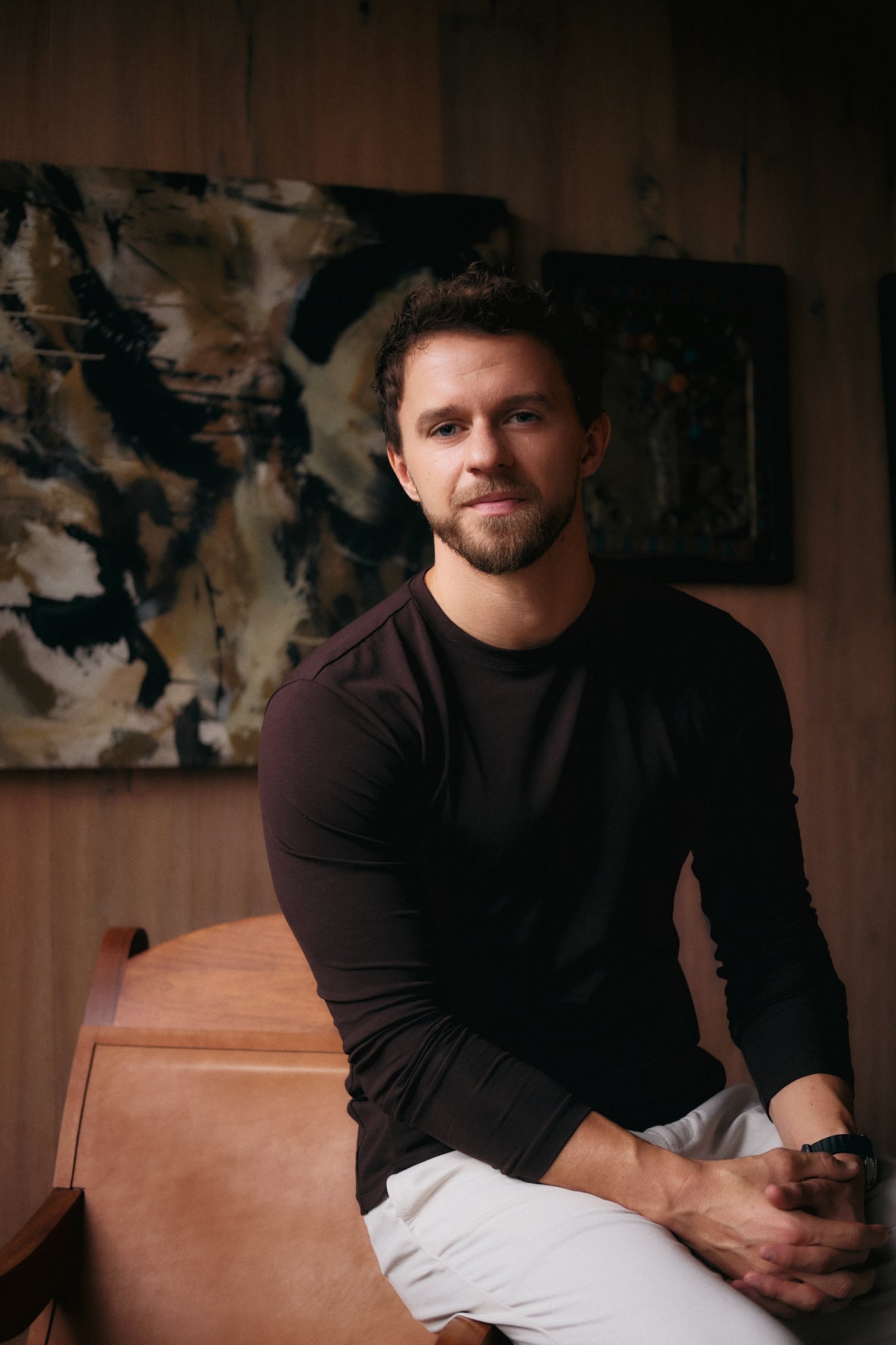 Editorial portrait of a young man with curly hair and short beard, wearing a dark long-sleeve shirt and white pants, seated casually on the edge of a wooden desk with hands clasped. He looks directly into camera with a relaxed, confident expression. 