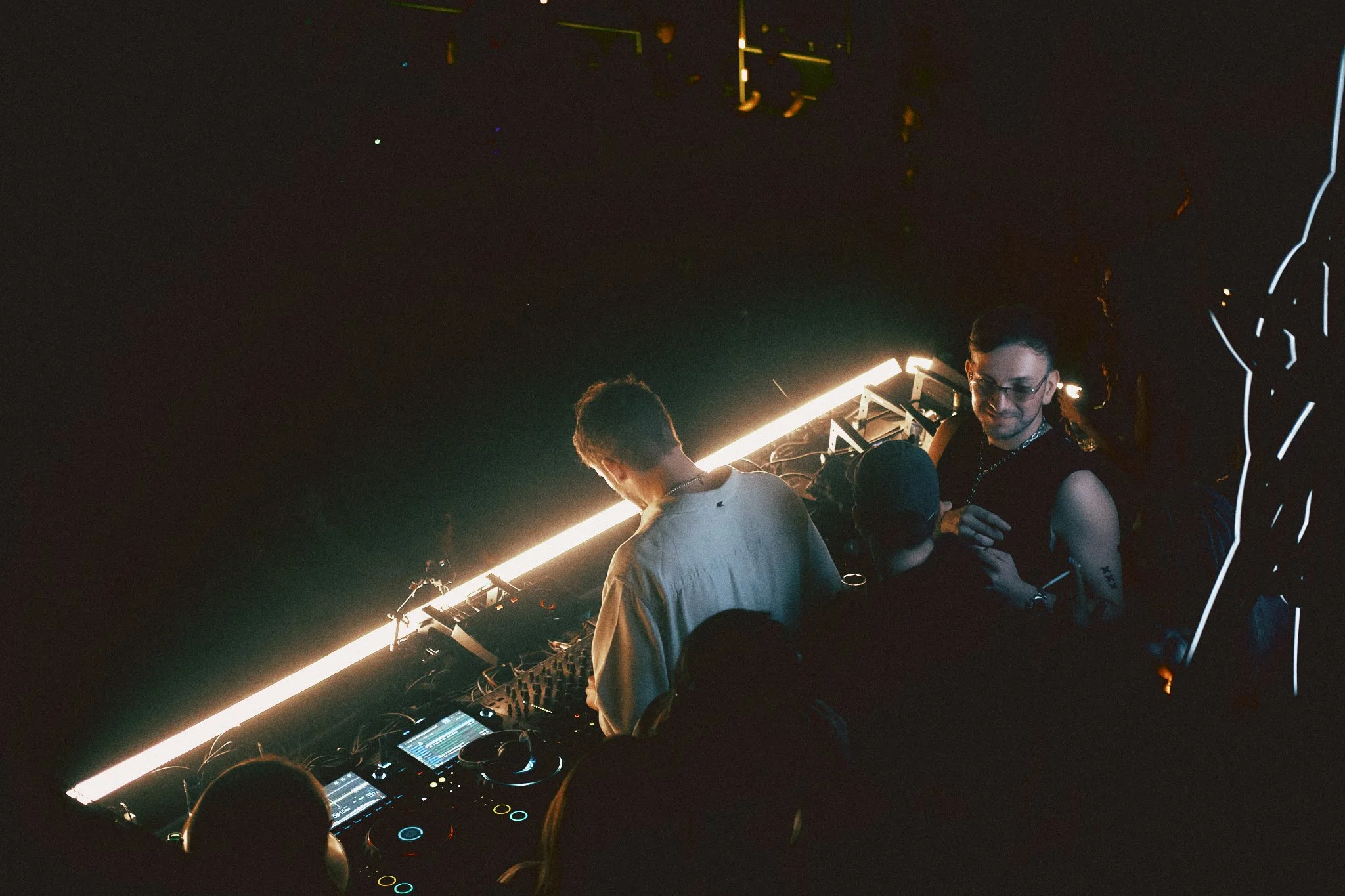 Two DJs in a dark nightclub, one wearing glasses and a sleeveless shirt, the other with a white shirt, surrounded by DJ equipment and people dancing.