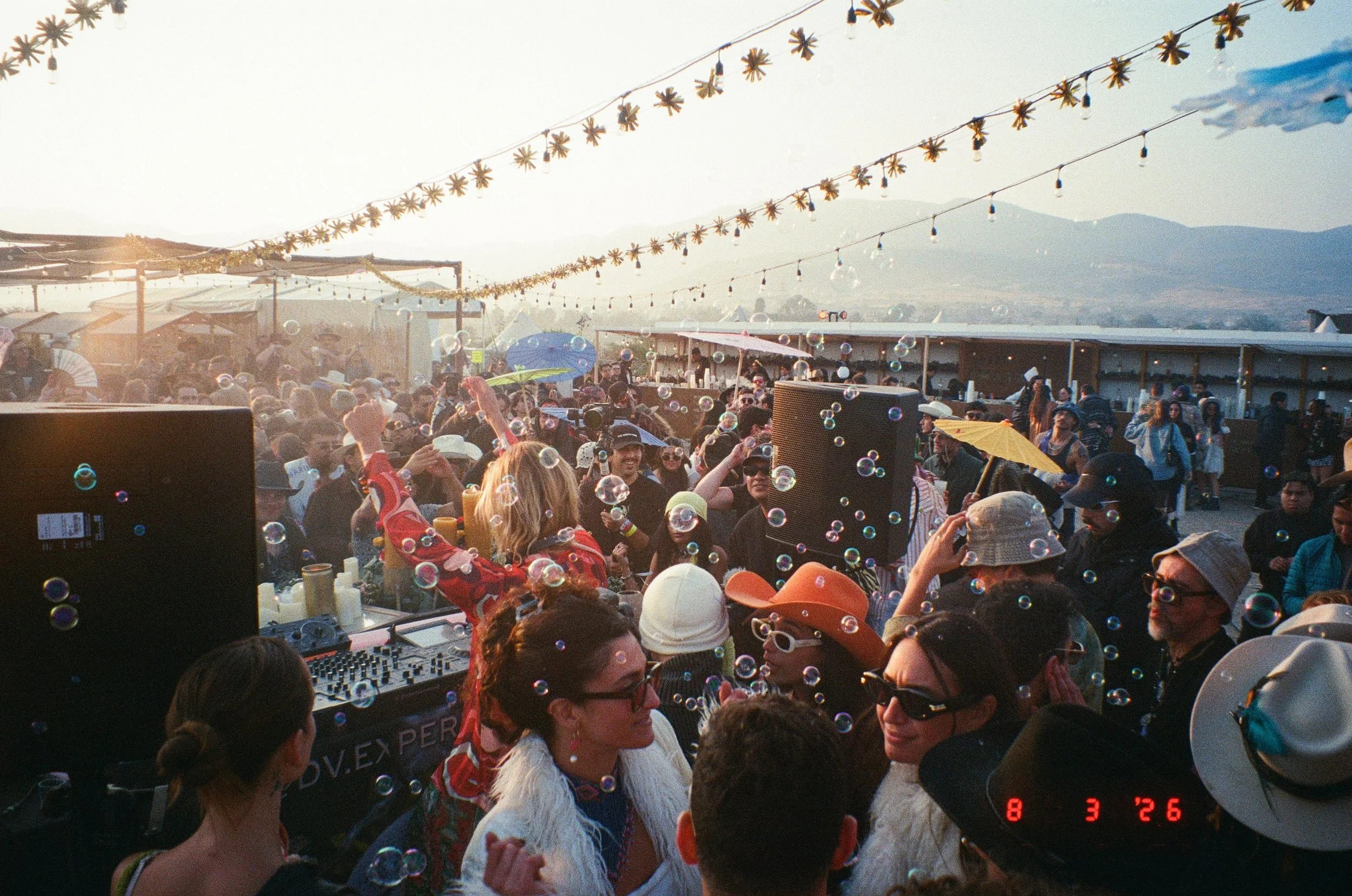 A large crowd of people gathered on the stage at Amarás Oaxaca Experience 2026 during sunrise, with decorative string lights and mountains in the background, as soap bubbles float through the air.